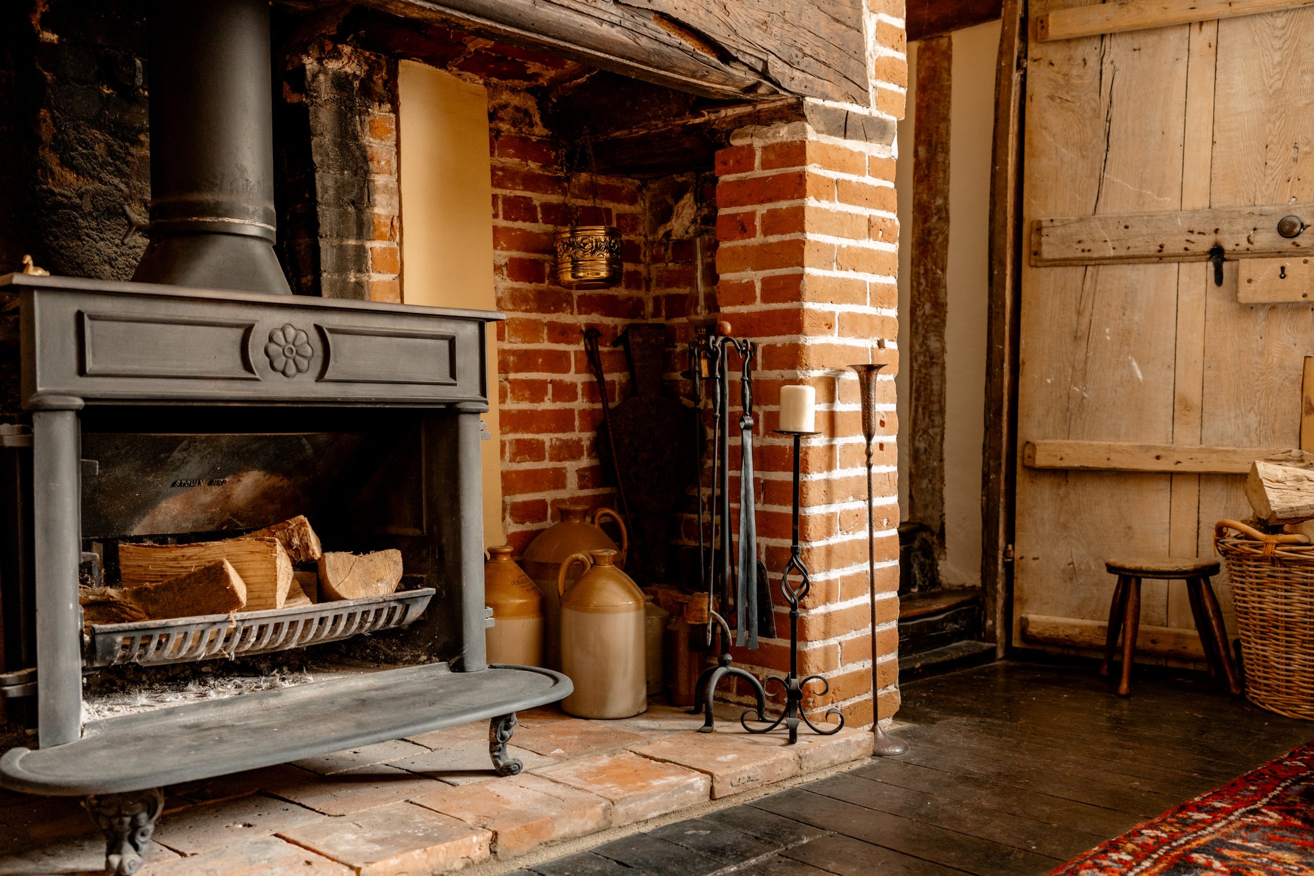 A rustic living area with a black wood-burning stove set into a red brick fireplace. Next to the stove are fire tools, candles, and ceramic jugs. The wooden floor has a small stool and a wicker basket. The wall features a distressed wooden door.