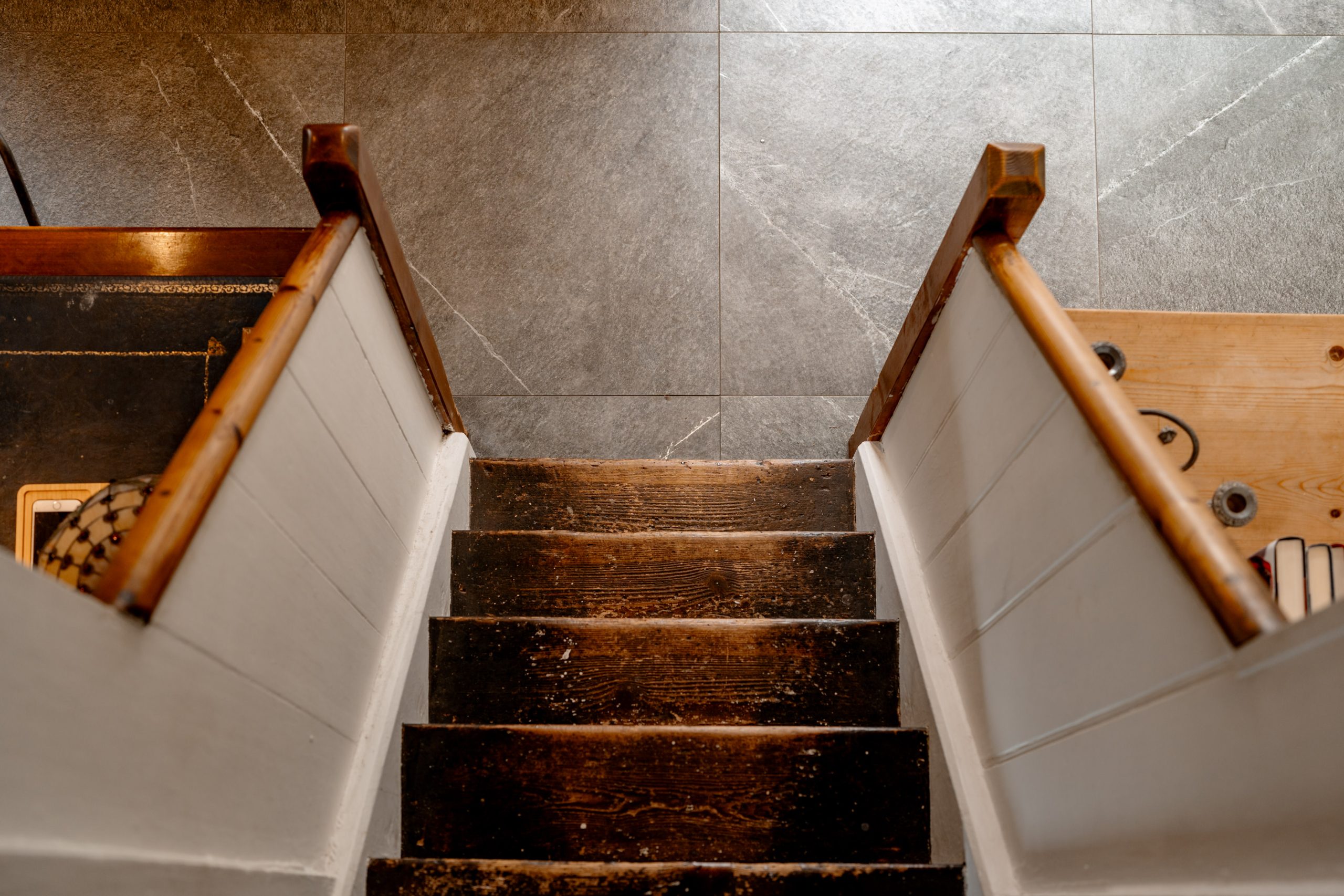 A downward view of a wooden staircase with white side panels and brown handrails leads to a tiled floor at the bottom. The worn steps display signs of frequent use.
