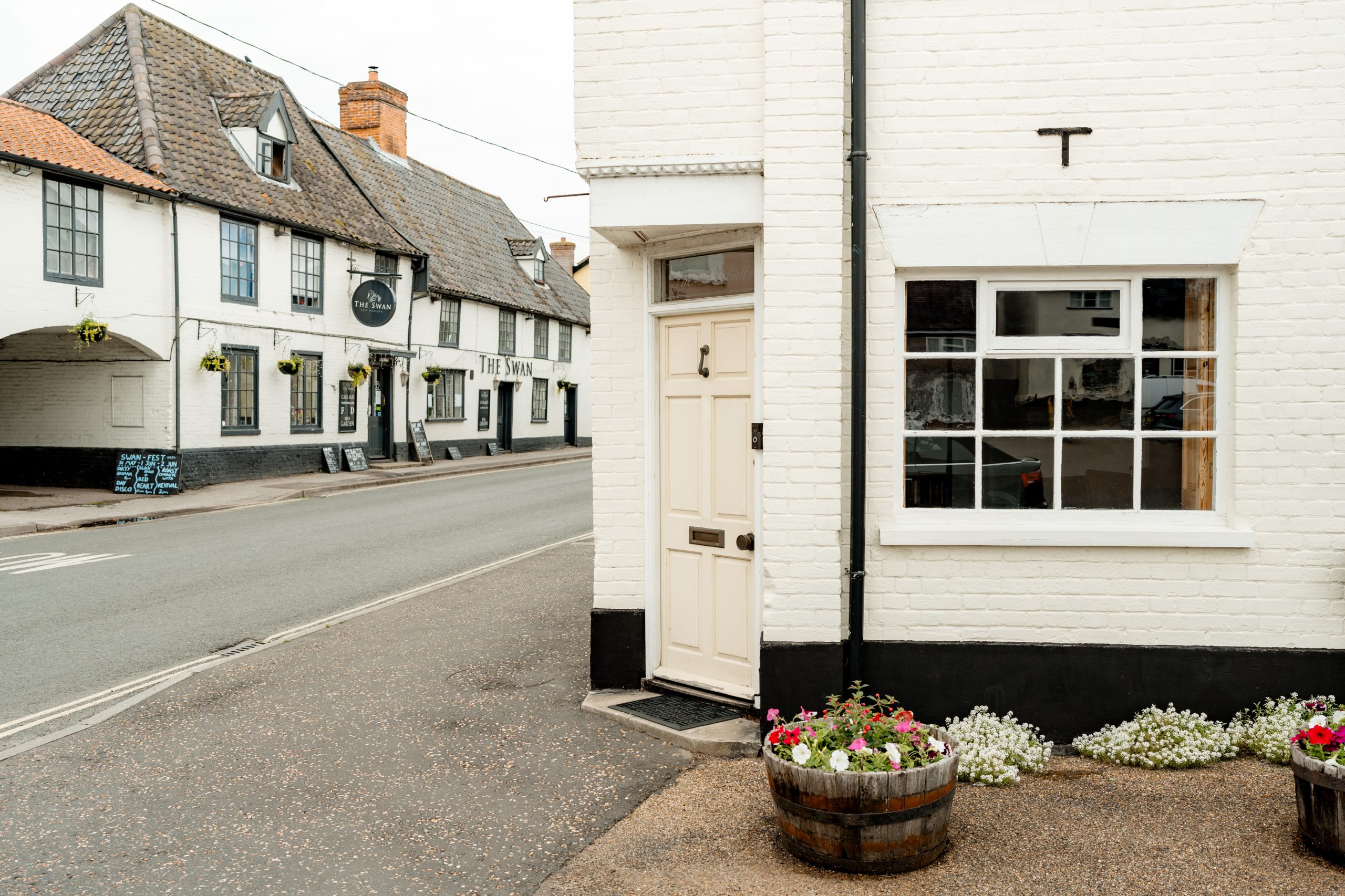 Street view of a quaint village with white brick buildings on either side. Visible are a door and window of a white building, flower barrels, and across the street, a building with