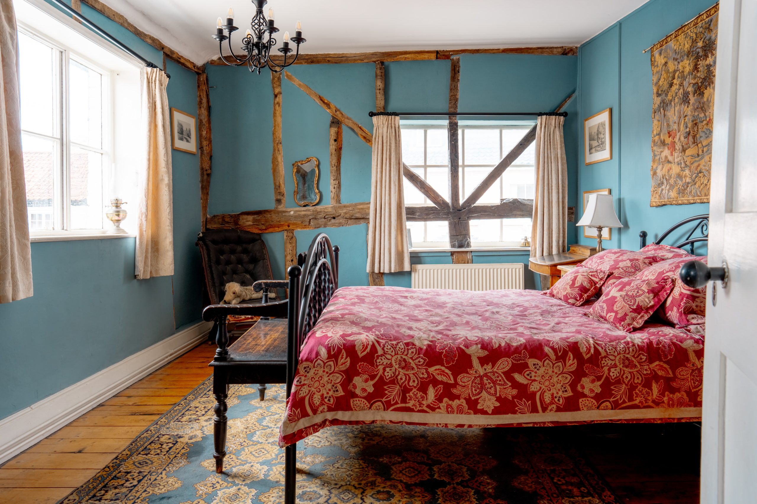 A cozy bedroom with teal walls and wooden beams features a double bed with red floral bedding. A black chandelier hangs from the ceiling, and a vintage armchair sits by the window. Light pours in through large windows with beige curtains, illuminating the room.