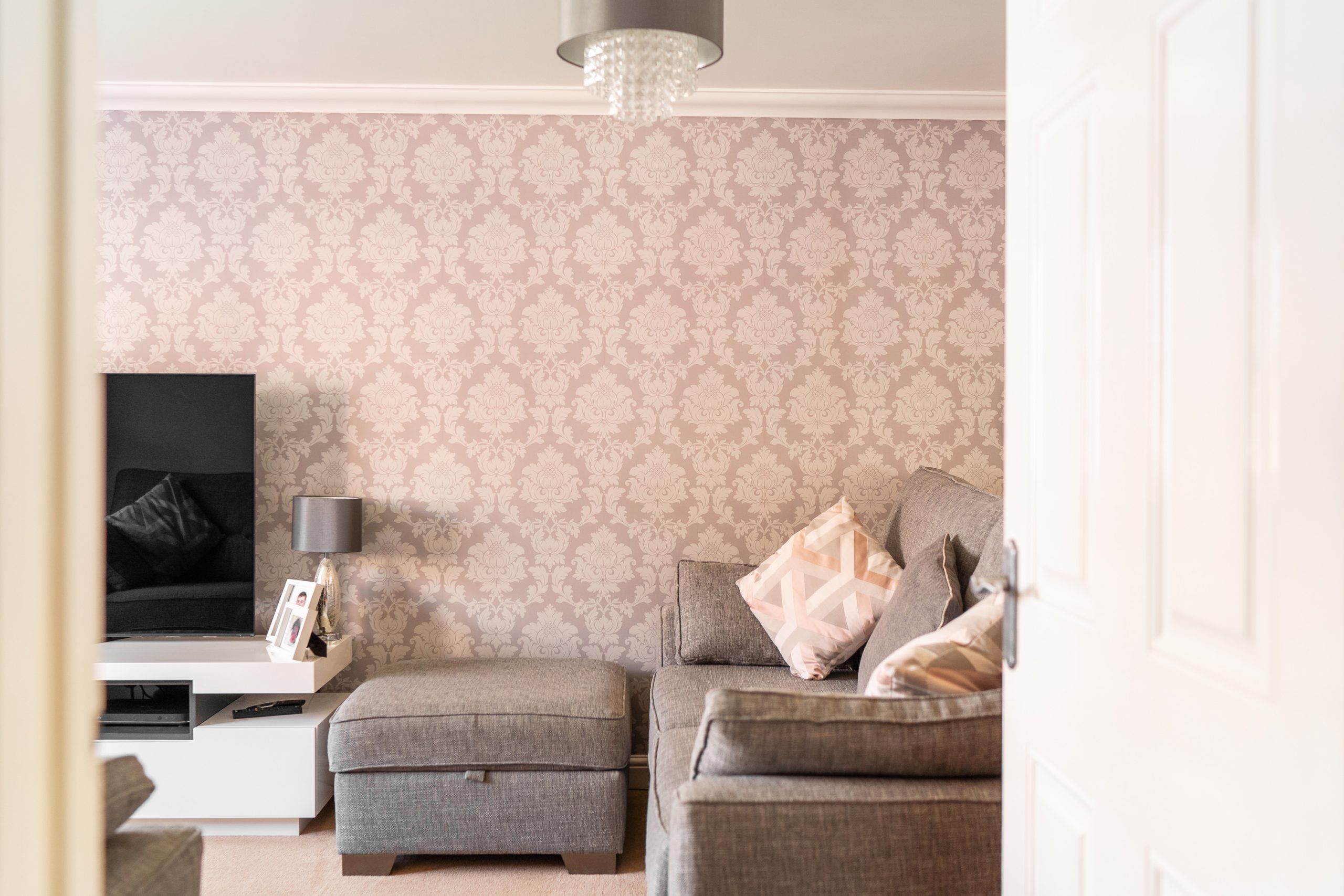A cozy living room with a gray sofa adorned with patterned cushions, an ottoman, and a white entertainment unit with a flat-screen TV. A small lamp sits on the unit. The walls are decorated with light pink floral wallpaper and a chandelier hangs from the ceiling.