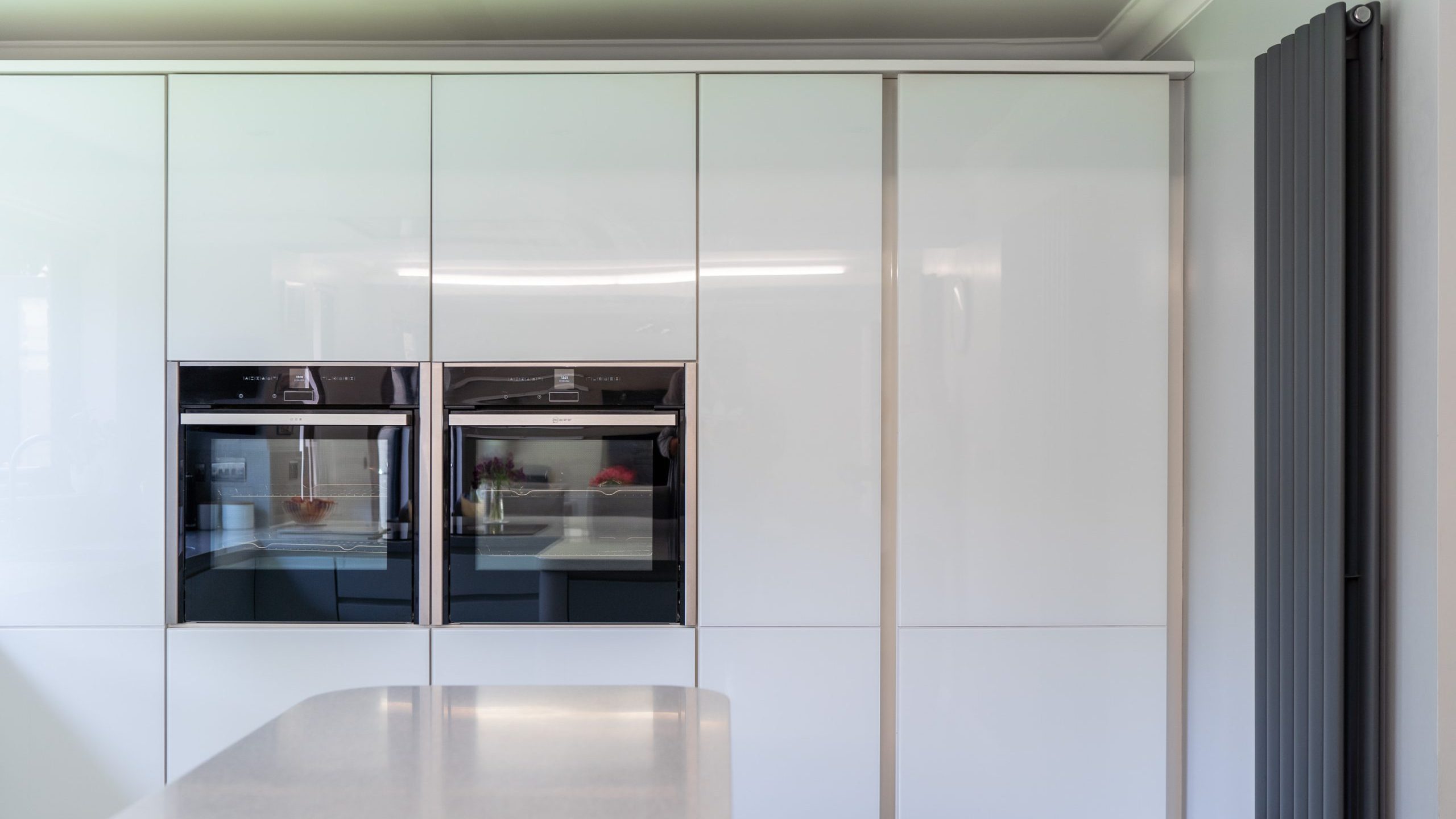A modern kitchen with sleek white cabinetry. Two built-in stainless steel ovens are aligned side by side within the cabinetry. The right side features a vertical radiator, and the foreground shows a white countertop with subtle reflections.