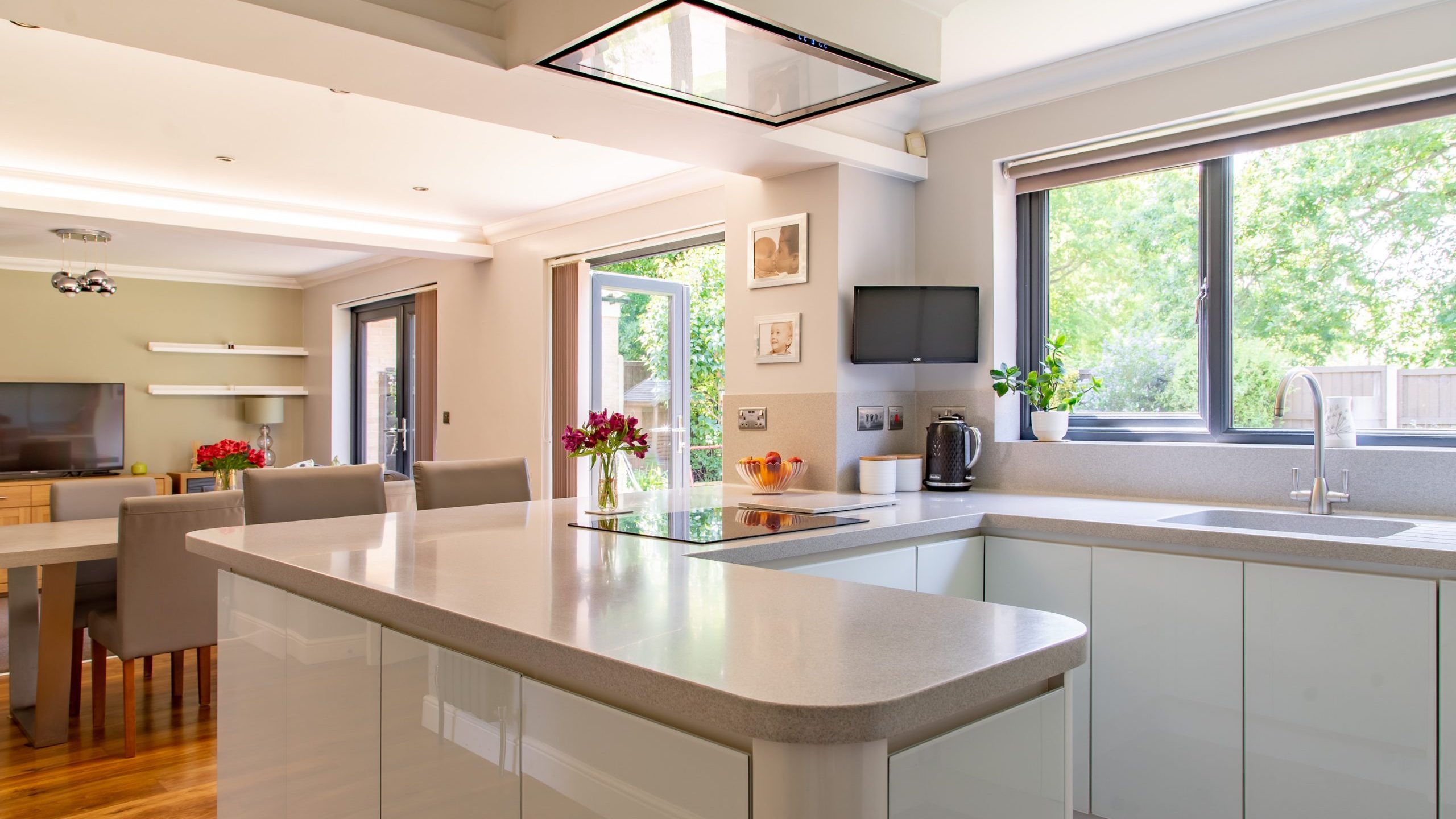 A spacious, modern kitchen with a large island countertop. It has white cabinets, a built-in stovetop on the island, and large windows letting in natural light. There is a dining area with a table and chairs in the background, decorated with fresh flowers.