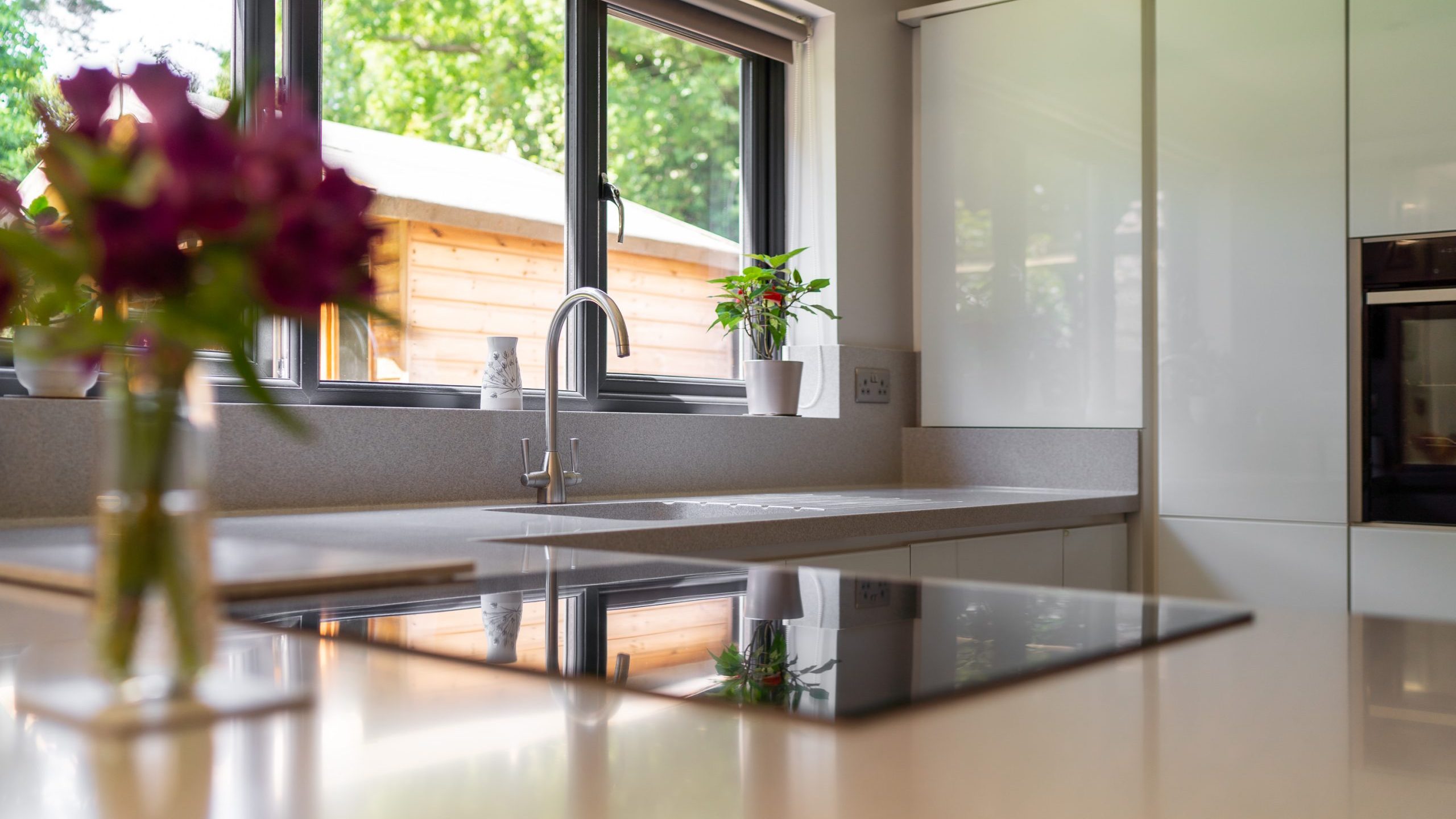 A modern kitchen with sleek white countertops and cabinets, large windows offering natural light and a view of a wooden shed outside. A stovetop and sink are installed on the countertop, with potted plants on the windowsill and a blurry bouquet in the foreground.