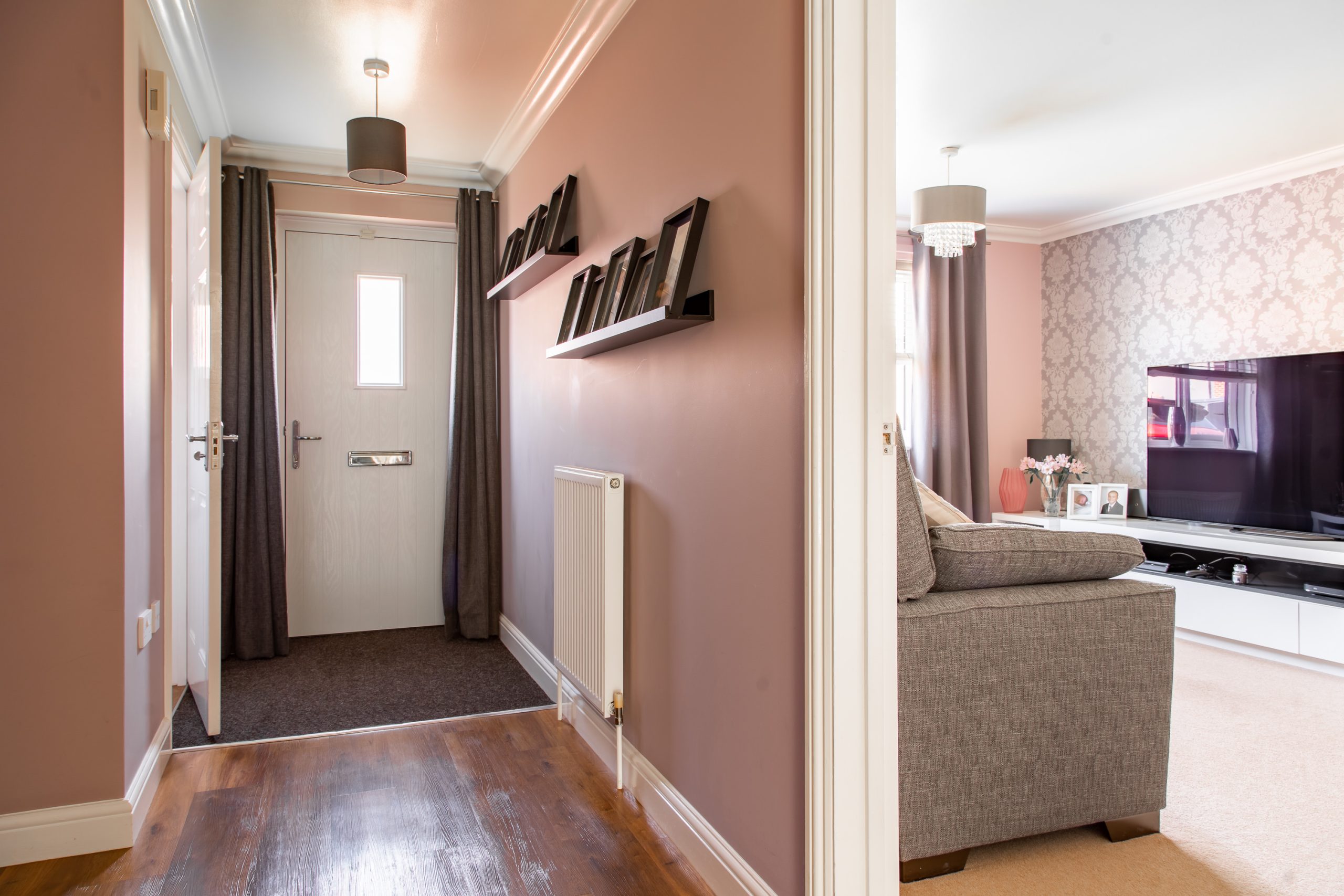 A modern home interior with a hallway and a living room. The hallway has wooden flooring, a white door, and a radiator. Shelves with decorative items hang on the wall. The living room to the right features a grey sofa, a large TV, and patterned wallpaper.