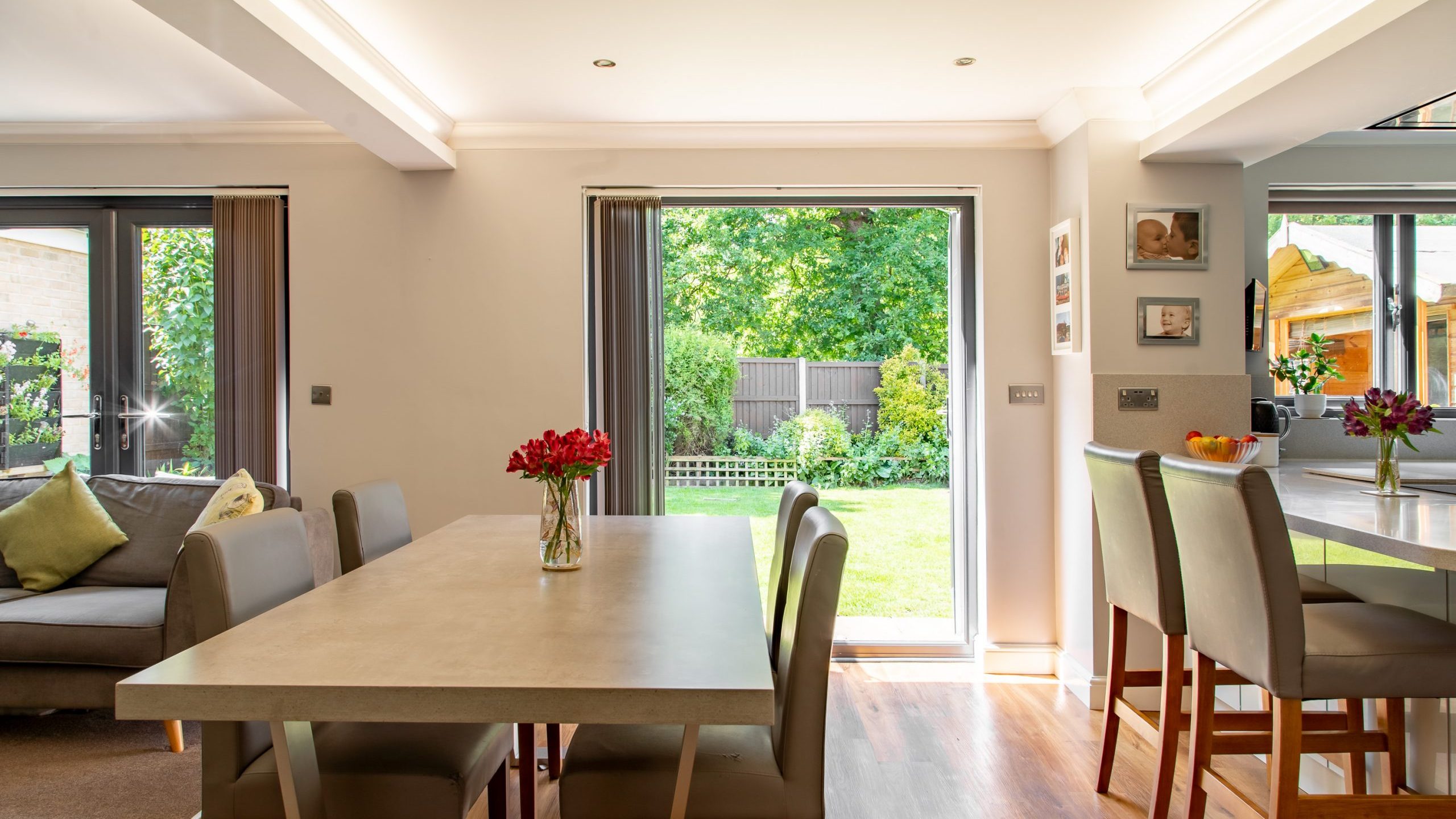 A bright, modern dining area with a table and six chairs, a small vase of flowers, and a bar counter with stools. Sliding glass doors open to a green garden with a wooden fence. The room features neutral tones with sunlight streaming in through the windows.