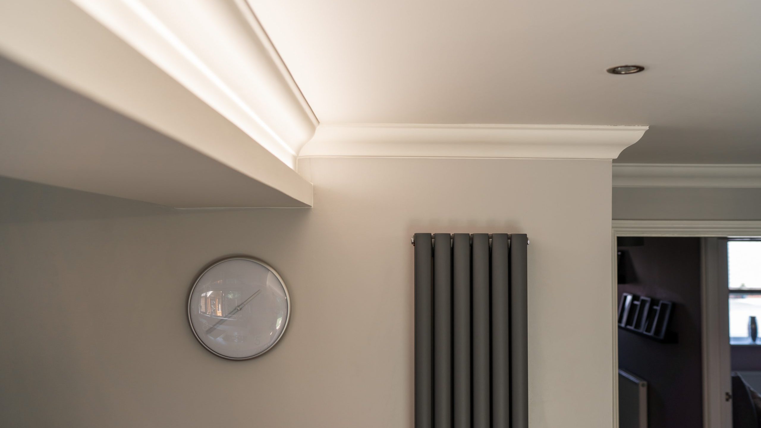 A modern interior with beige walls, featuring a white circular wall clock and vertical dark grey radiator. Molding is visible along the ceiling edge. An open doorway shows a glimpse of another room with framed pictures on the wall and light from a window.