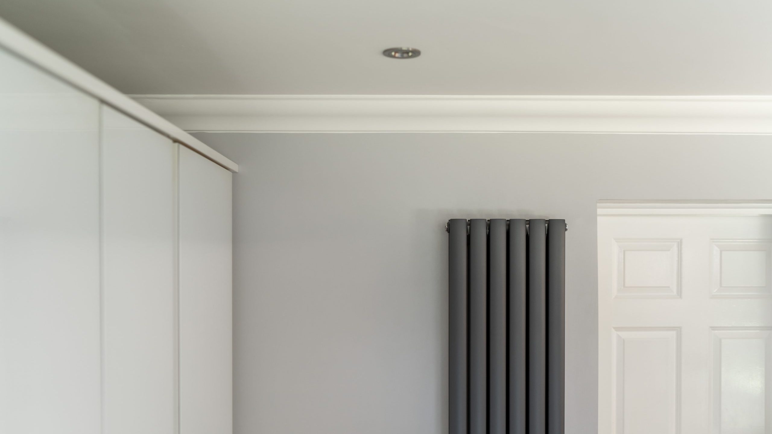 A minimalistic interior featuring a section of a white wardrobe on the left, a grey vertical radiator on a light grey wall in the center, and a partially visible white door on the right under a plain ceiling with a small recessed light.