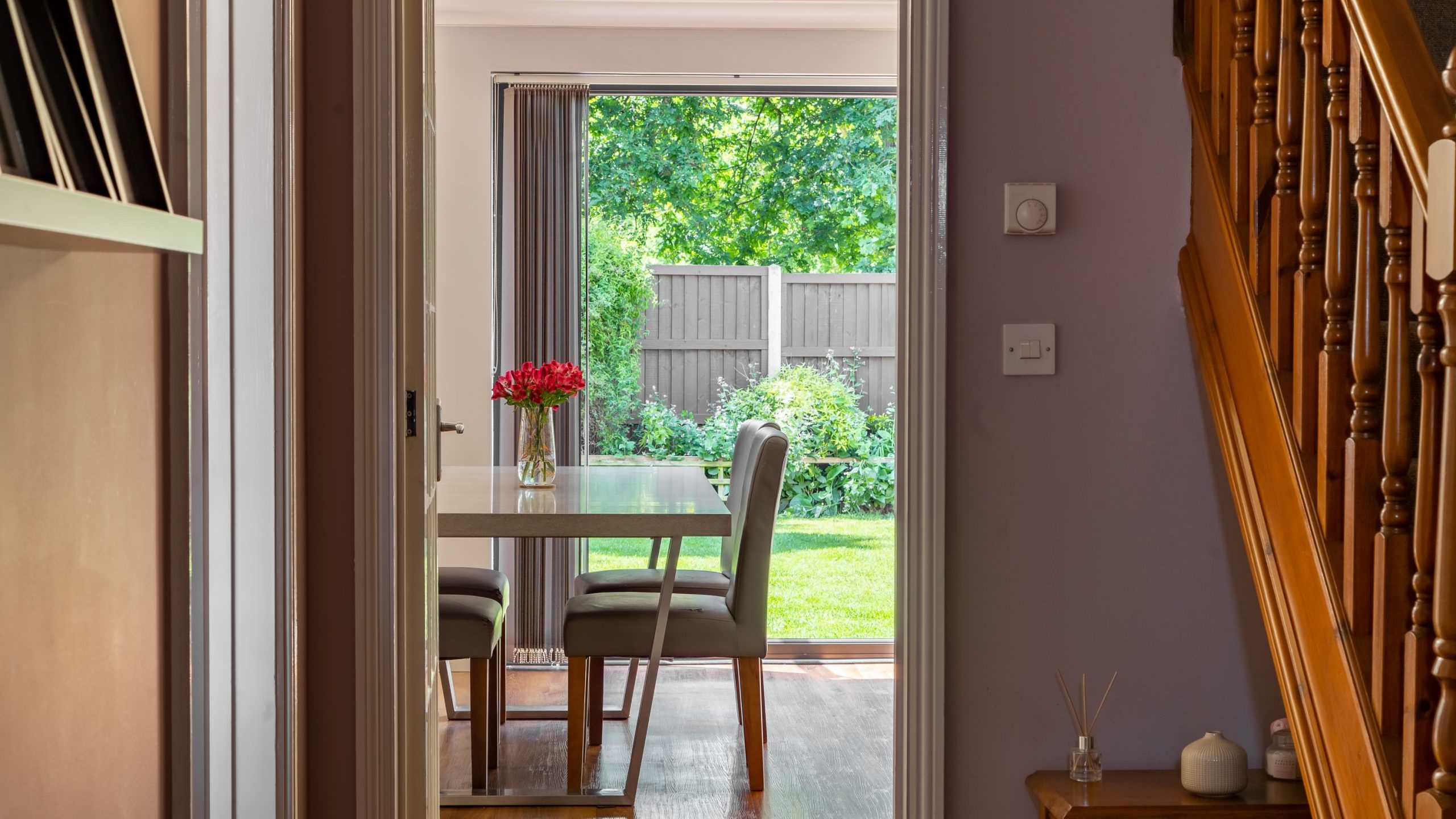 View through a hallway towards a dining area with a table and chairs. A vase of red flowers is on the table. The room has glass doors leading to a garden with green foliage and a wooden fence. Wooden stairs and a small table with candles are on the right.