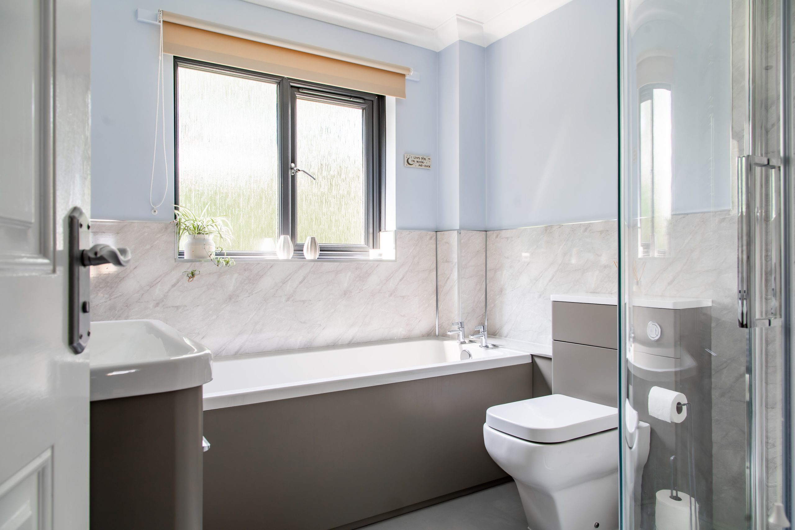 Bright, modern bathroom with a large window, bathtub, sink, toilet, and glass-enclosed shower. The walls are a light blue with marble accents. A small plant and toiletries are visible on the windowsill. The room is clean and well-lit with natural light.