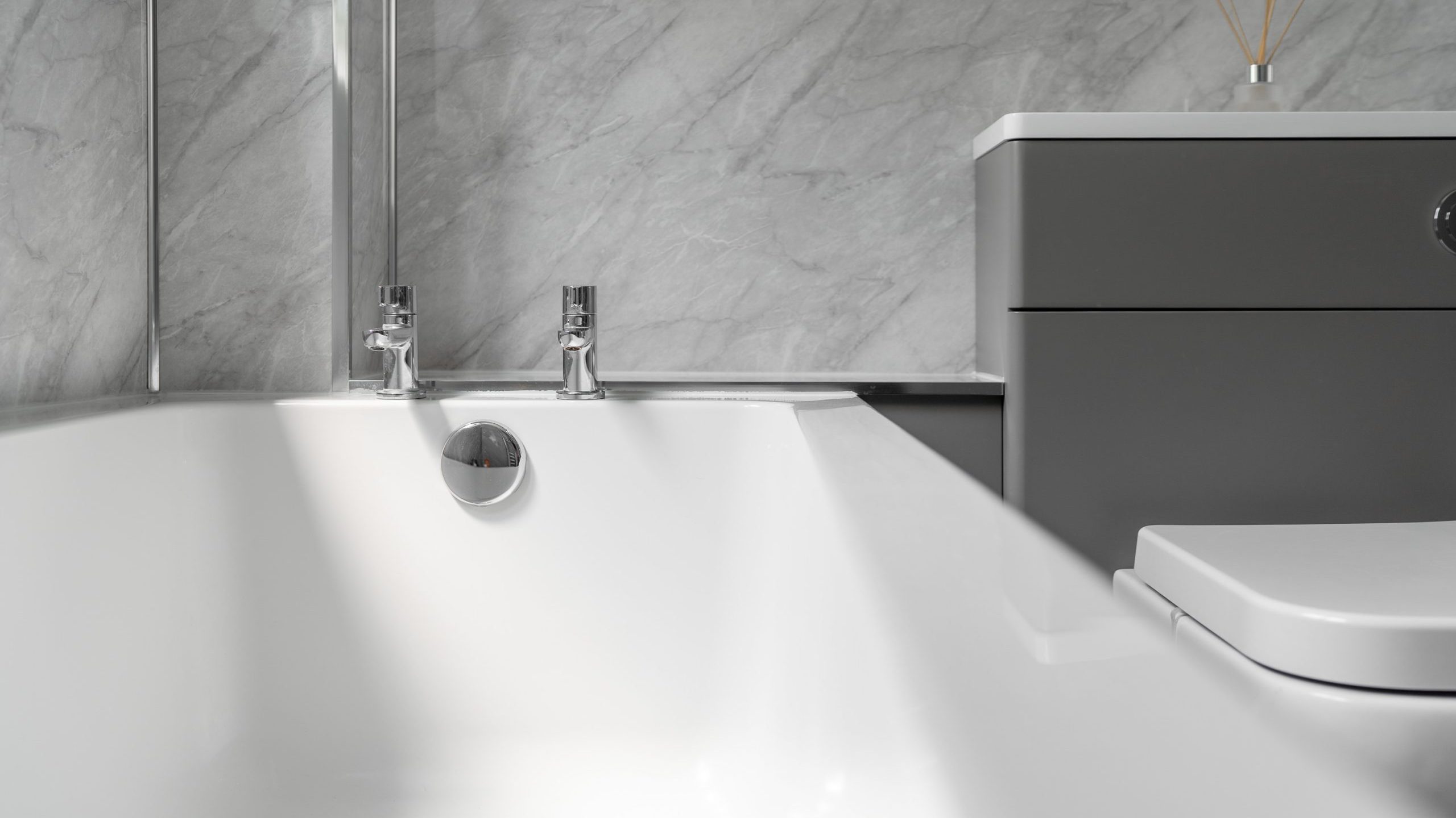 A contemporary bathroom with a white bathtub featuring chrome taps and drain. Adjacent is a modern gray vanity with a countertop adorned with a minimalist reed diffuser. The backdrop is a sleek gray marble wall.