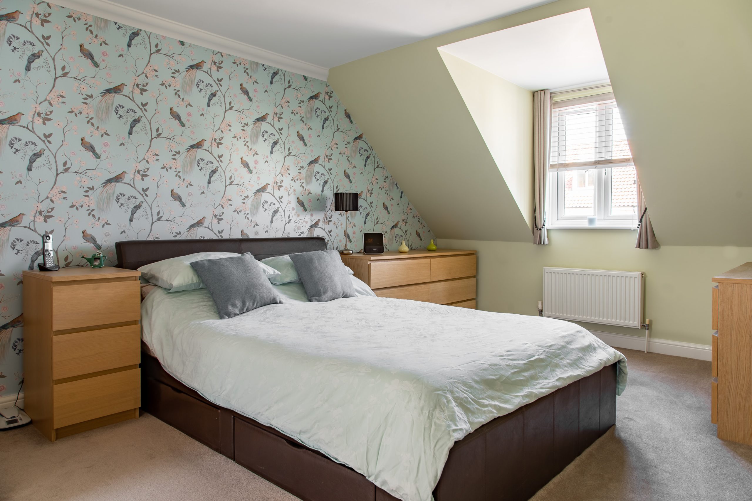 A cozy bedroom featuring a double bed with a light green comforter and gray pillows. A wallpapered accent wall with a bird pattern stands behind the bed. The room also has two wooden nightstands with lamps, a chest of drawers, and a window with blinds.