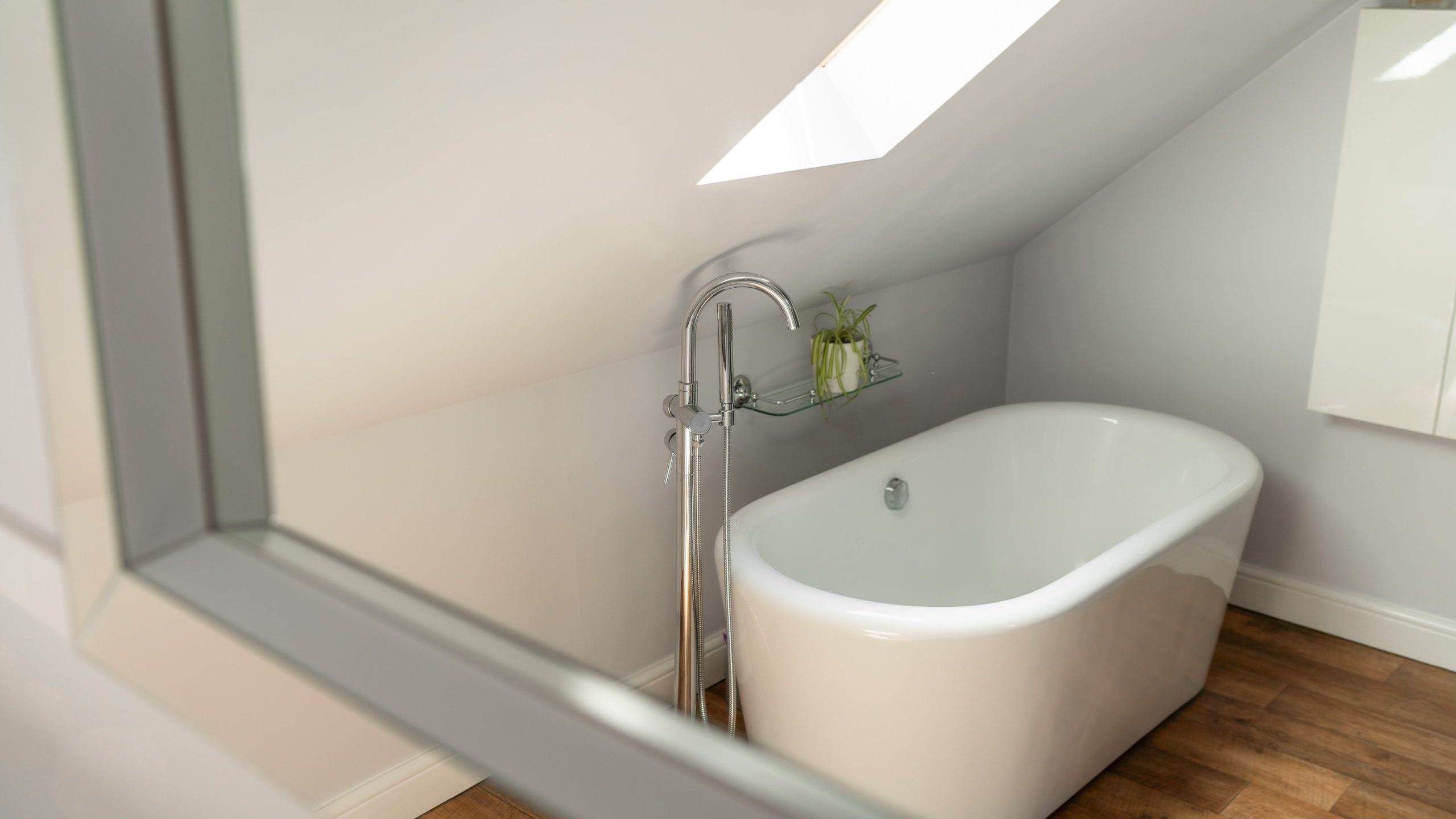 An attic bathroom features a white freestanding bathtub placed under a slanted ceiling with a skylight. The wooden floor adds warmth, and a small green plant sits on a glass shelf by the bathtub, creating a cozy, light-filled space.