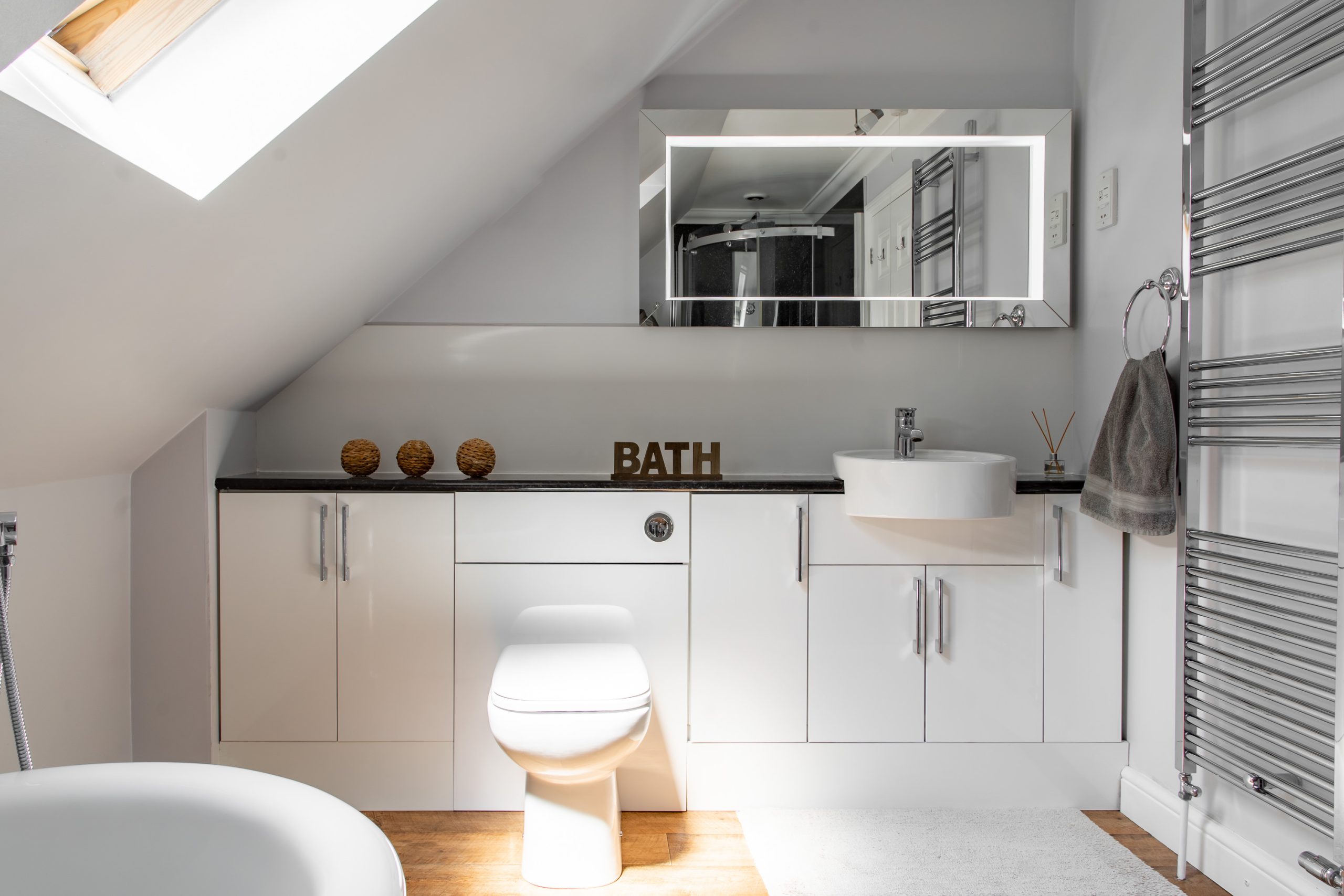 A modern white bathroom with a sloped ceiling, featuring a skylight, wall-mounted sink, and a toilet. There's a large illuminated mirror above the sink, decorative items on the countertop, and a towel hanging on a chrome radiator. The word