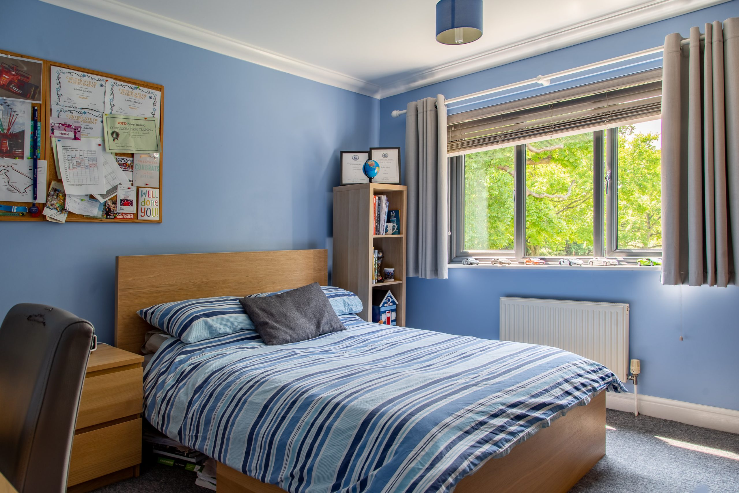 A neatly organized bedroom with blue walls. The bed has a striped blue, white, and gray comforter, and there's a wooden headboard. A corkboard filled with notes and photos hangs on the wall. A window with gray curtains offers a view of lush greenery outside.