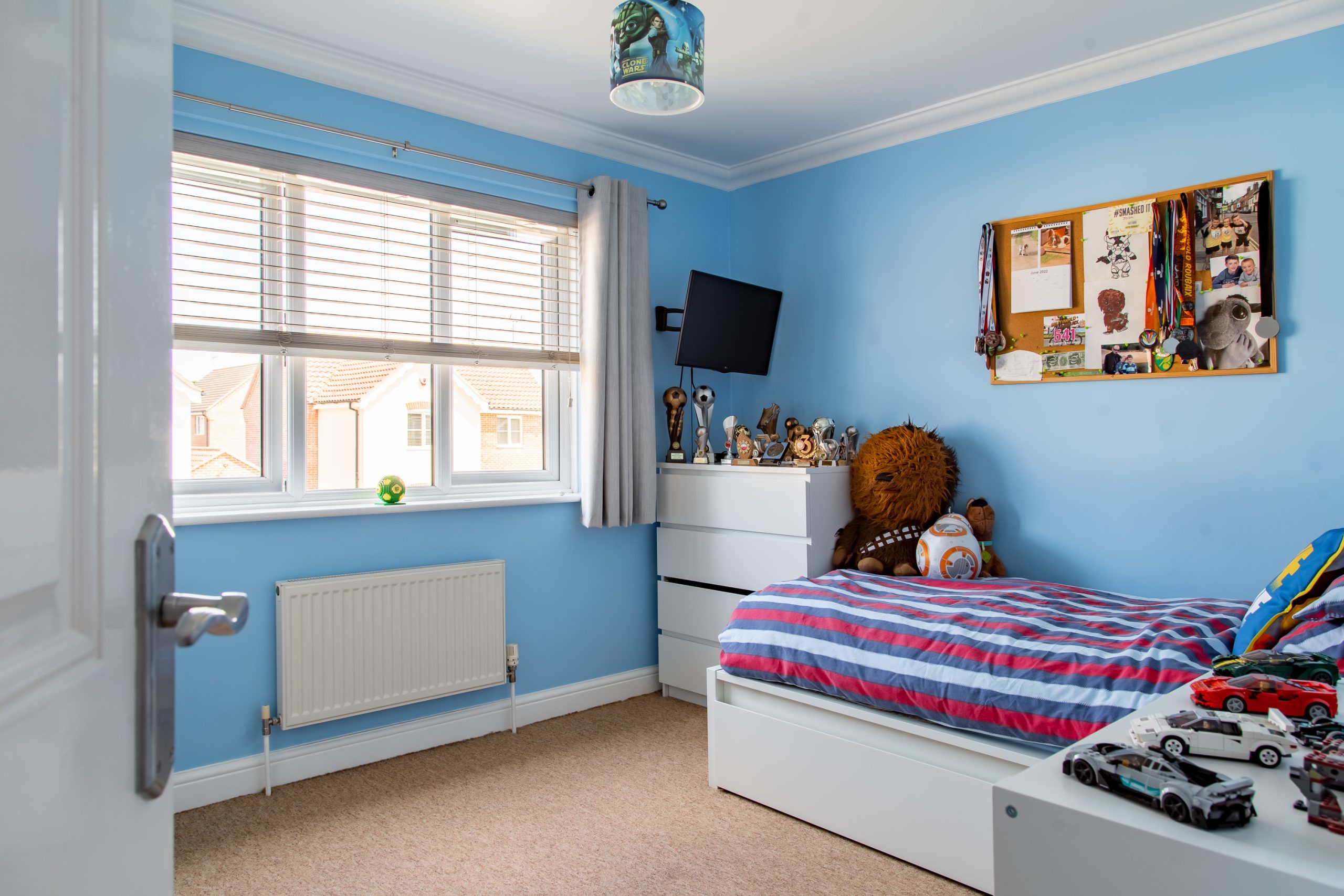 A neatly organized child's bedroom with blue walls, a striped bed, and a chest of drawers holding trophies and figurines. There's a bulletin board with pictures above the chest, a small TV mounted on the wall, and a toy lion on the bed. The window lets in natural light.