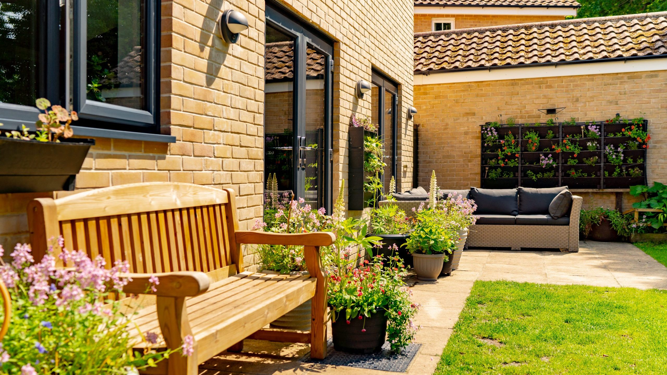 A sunny backyard patio features a wooden bench and various potted plants with blooming flowers. On the right, a cushioned outdoor sofa sits in front of a vertical garden wall with more plants. The area is surrounded by a brick house and lush green lawn.