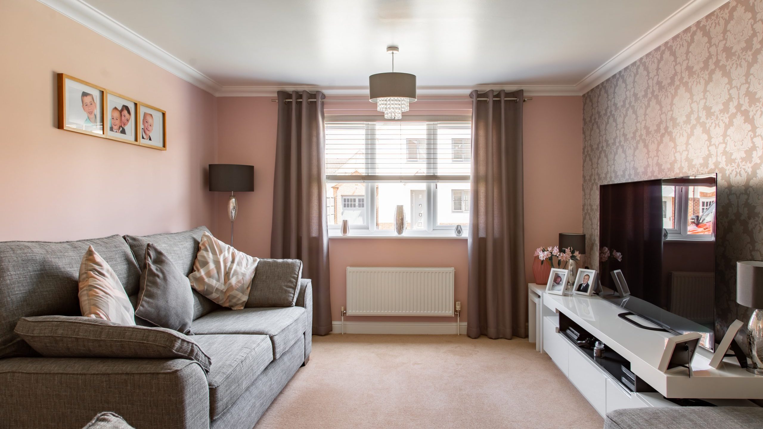 A cozy living room with light pink walls features a grey couch adorned with patterned cushions. Family photos are displayed on the wall and a TV sits on a white entertainment unit surrounded by more framed photos. Sunlight streams through the window with taupe curtains.