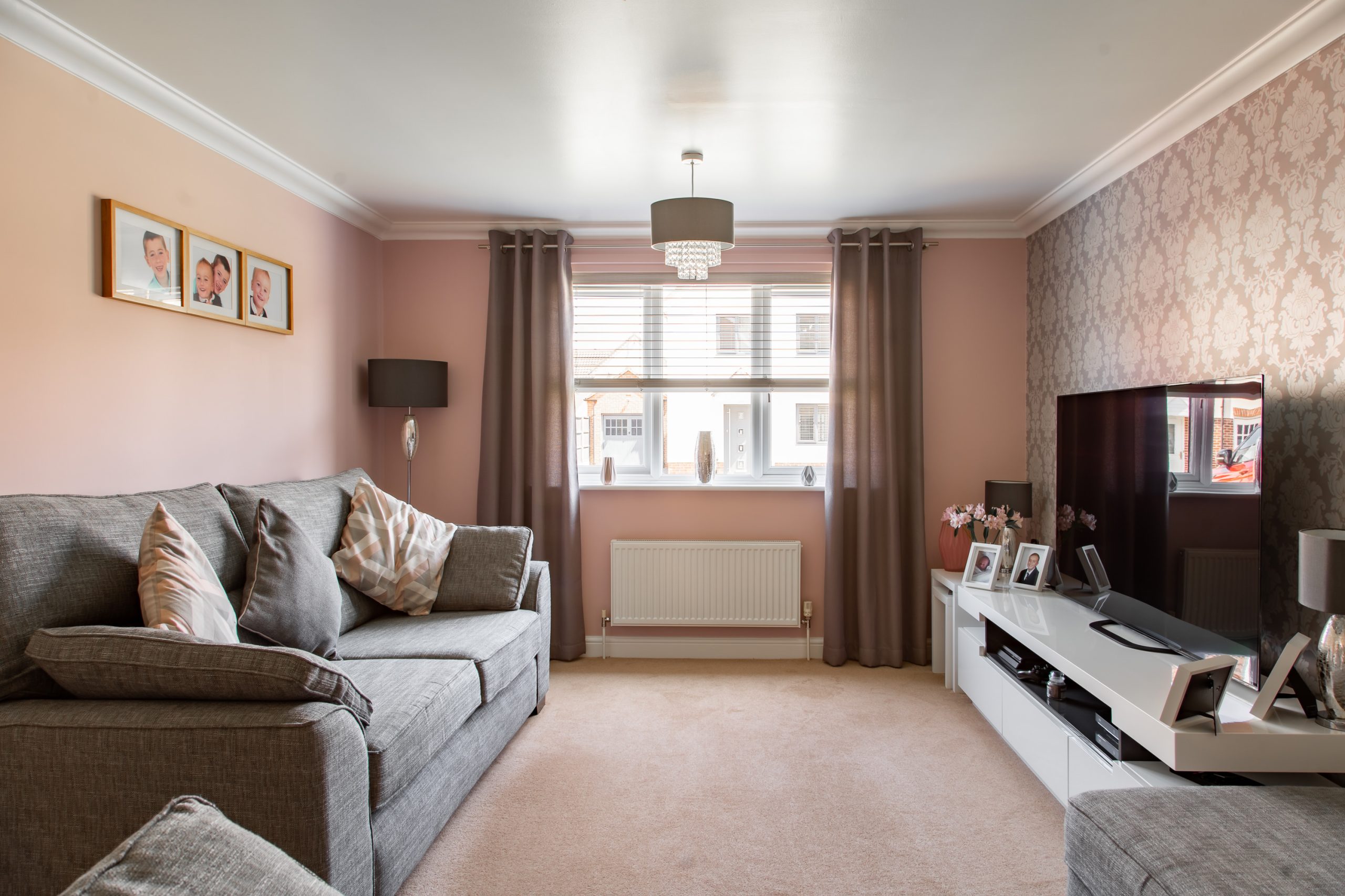 A cozy living room with light pink walls features a grey couch adorned with patterned cushions. Family photos are displayed on the wall and a TV sits on a white entertainment unit surrounded by more framed photos. Sunlight streams through the window with taupe curtains.