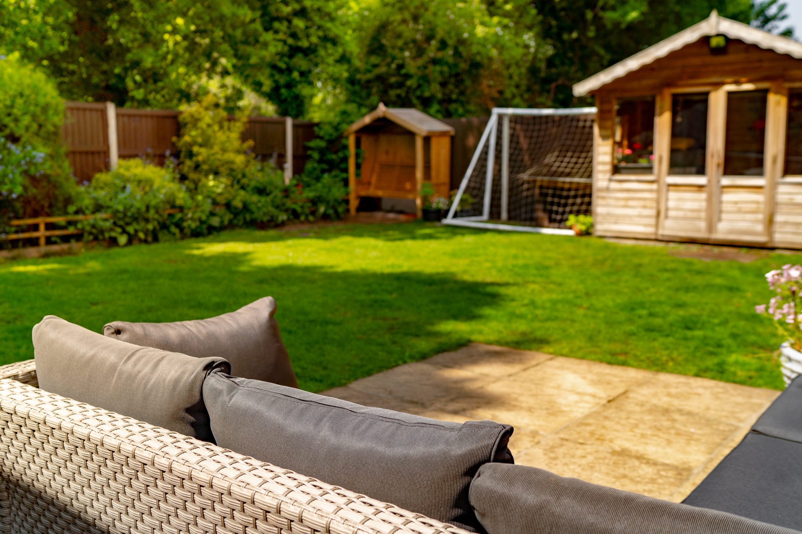A cozy backyard with a wicker sofa topped with gray cushions in the foreground. In the background, there is a lush green lawn, a small wooden shed, a wooden playhouse, and a soccer goal. Fenced garden area with trees and bushes surrounding. Bright and sunny day.