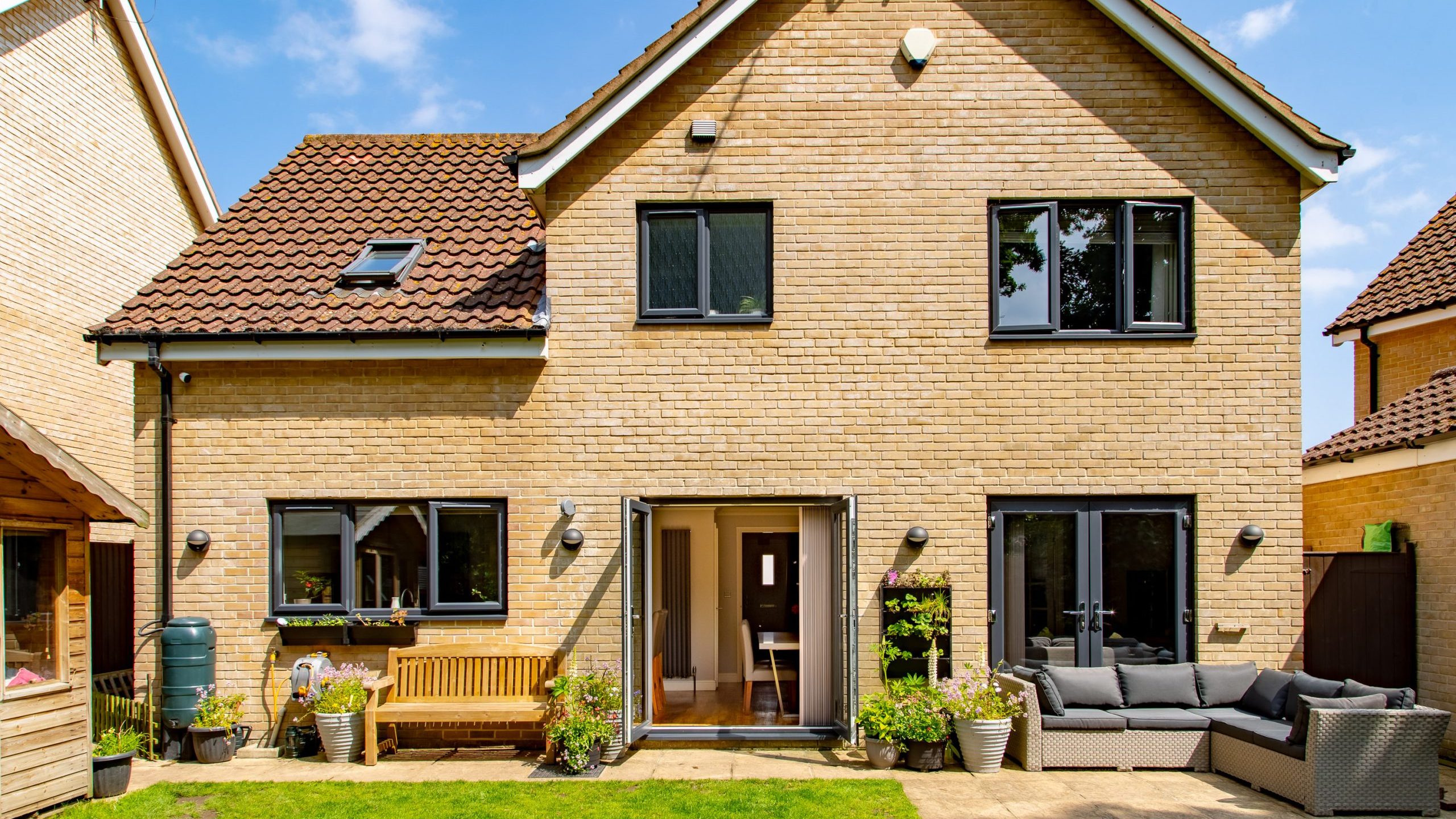 A two-story house with light brick exterior, featuring a red-tiled roof and multiple windows. The backyard has a neatly maintained lawn, potted plants, a wooden bench, and a gray outdoor sofa set on a patio. A sunny day with blue sky and scattered clouds.