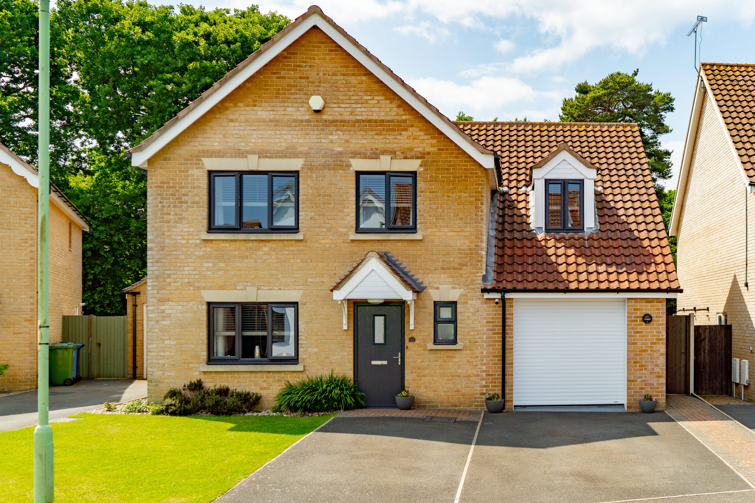 A two-story detached house with a brick exterior and gable roof. It features a grey front door, double-pane windows, and a garage with a white roll-up door on the right. The front yard has a well-maintained lawn, a driveway, and a green trash bin on the left.