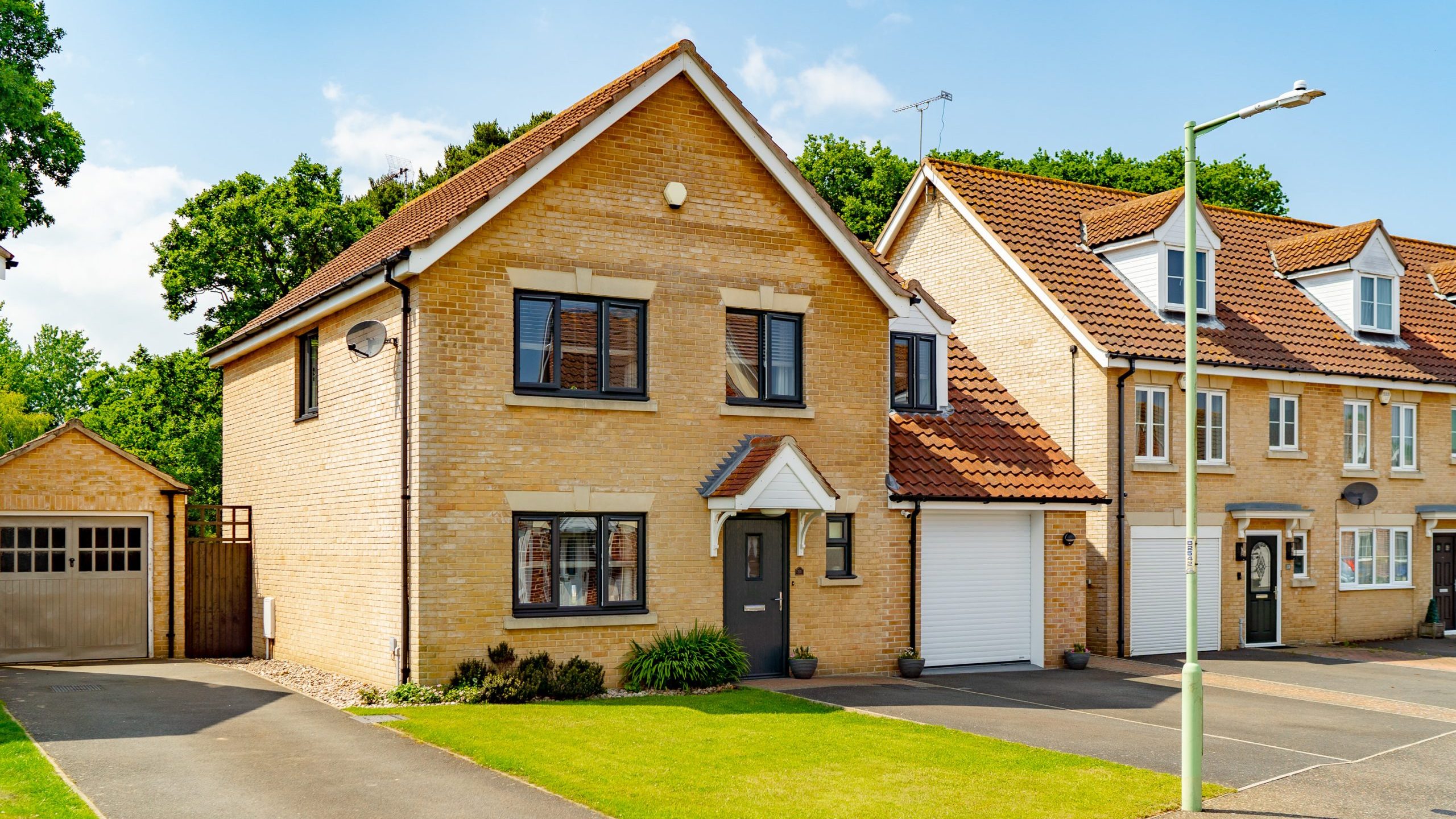 A detached, two-story brick house with a pitched roof, a small front garden, and a driveway. The house has white-framed windows and a dark front door with a small overhang. Beside it is an attached garage. Neighboring houses are seen on the right.