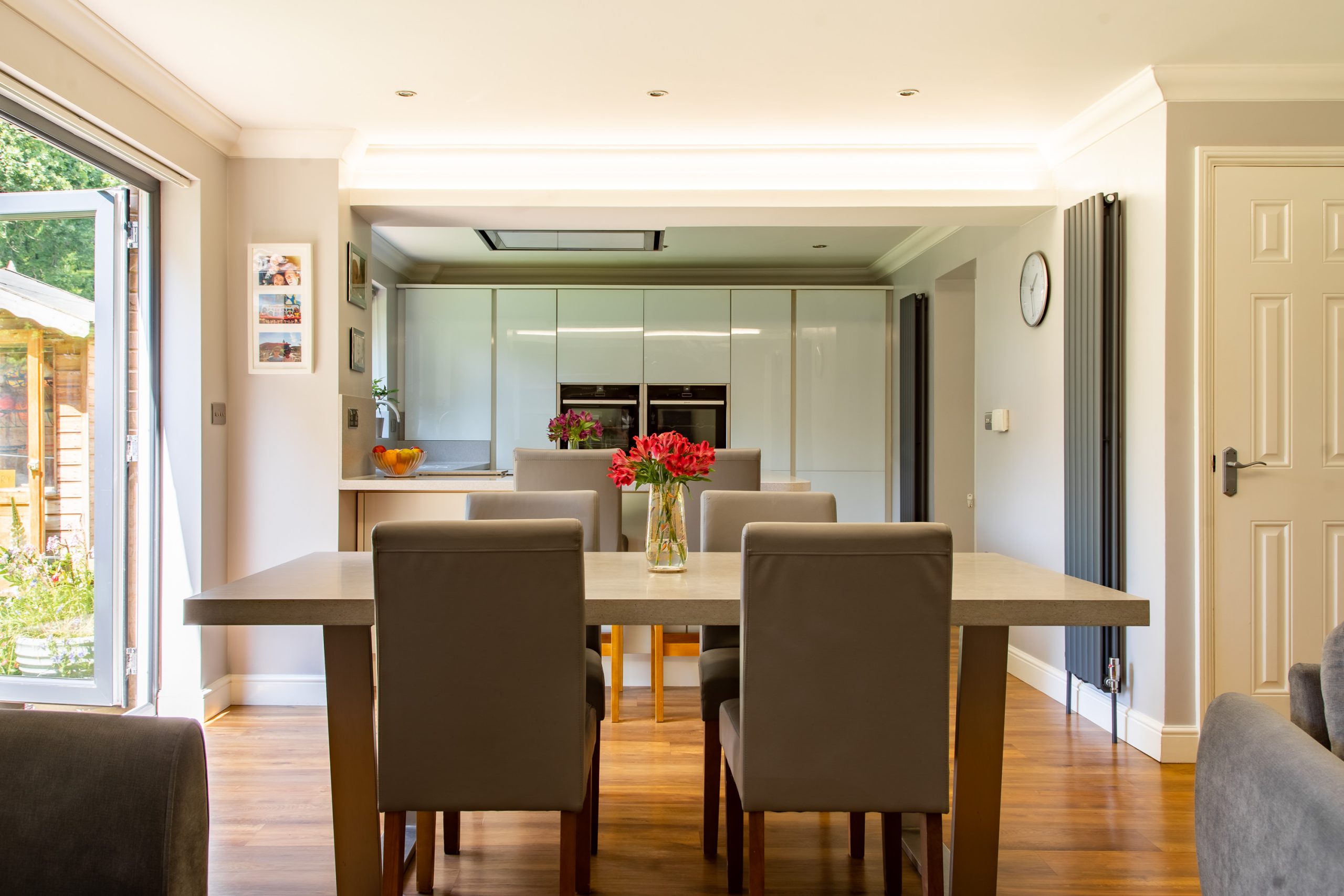 A modern kitchen and dining area featuring a wooden dining table with six gray cushioned chairs. A vase with red flowers sits atop the table. The kitchen has sleek white cabinets and built-in ovens. A door to the left leads outside, letting in natural light.