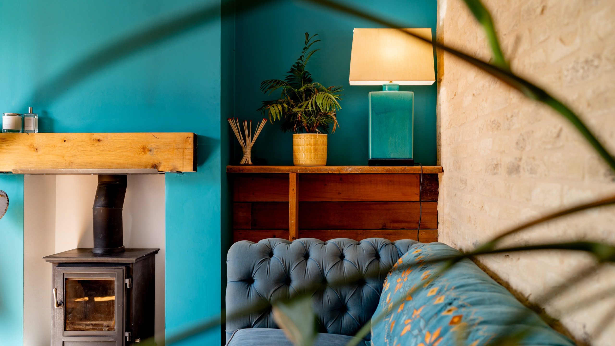 A cozy living room with teal walls and a light brick accent wall. A black wood-burning stove is set against the teal wall, and a tufted teal sofa sits nearby. A wooden shelf holds a potted plant and a teal lamp with a beige shade. Some leaves are in the foreground.