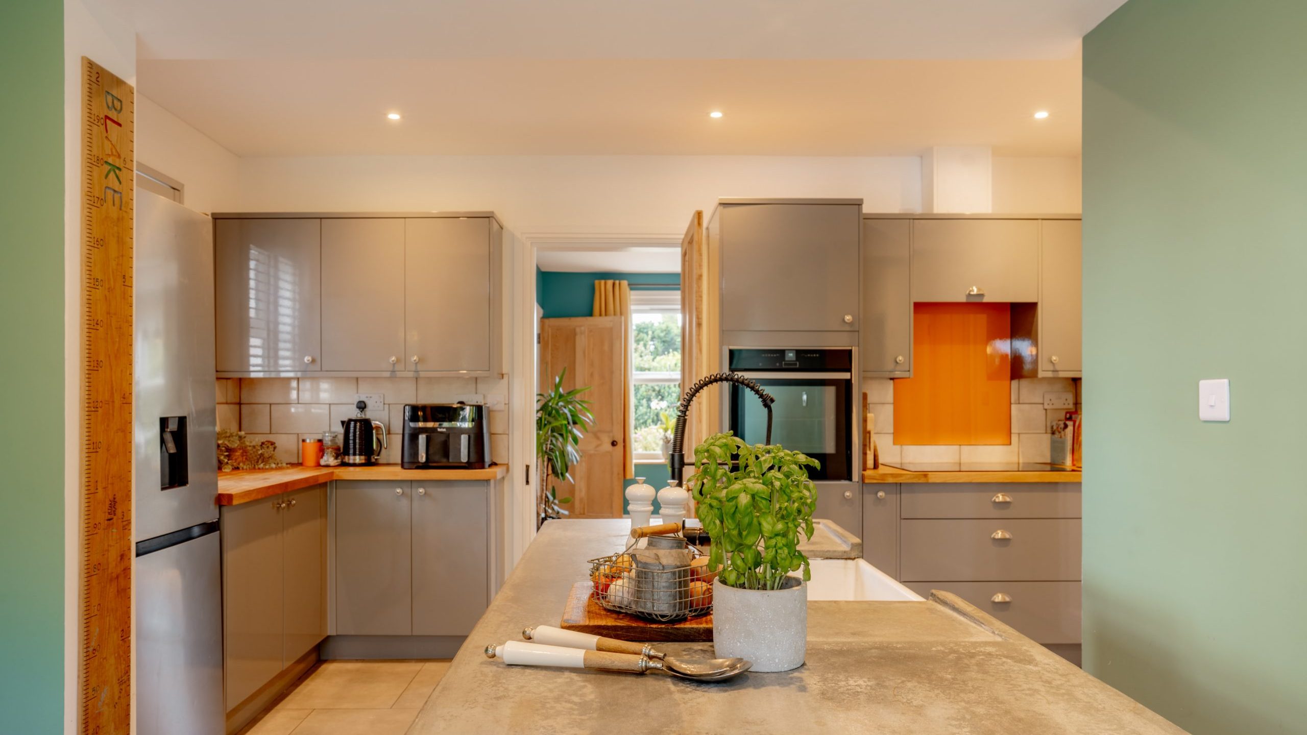 A modern kitchen with green walls, beige cabinets, and stainless steel appliances, including a fridge and oven. A large island in the foreground has a faucet, potted plant, and wooden cutting board with kitchen utensils. A doorway and window can be seen in the background.