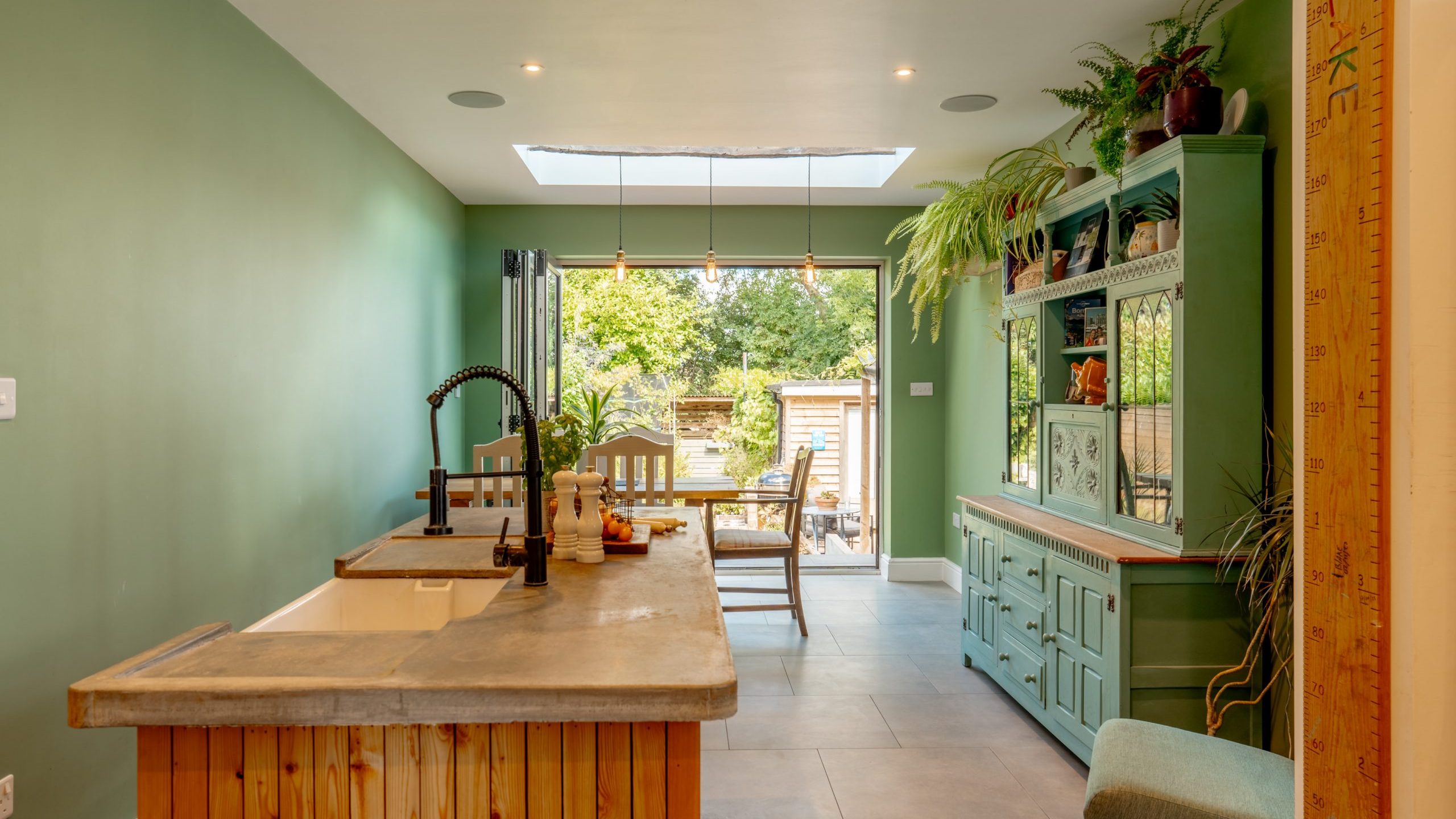A cozy kitchen with green walls features a wooden island with a concrete countertop, a black faucet, and a farmhouse sink. Behind it is a rustic green cabinet adorned with plants. Large windows and a glass door provide a view of a garden with outdoor seating.