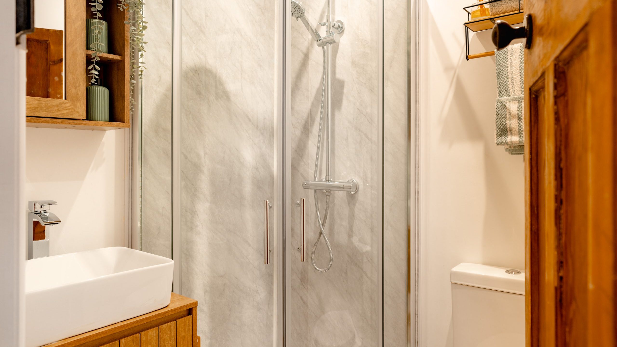 A small bathroom features a modern design with a glass-enclosed shower, a white ceramic sink on a wooden vanity, and a minimalistic toilet. Wooden cabinet and shelves with green decor accentuate the space, providing a cozy and stylish atmosphere.