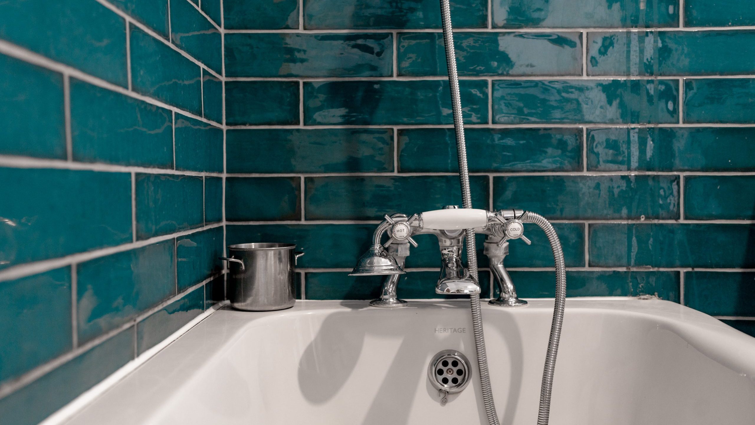 A white bathtub with a vintage-style chrome faucet and handheld showerhead is set against teal subway tile walls. A small metal bucket sits on the edge of the tub. The tiles have a glossy finish, reflecting light.
