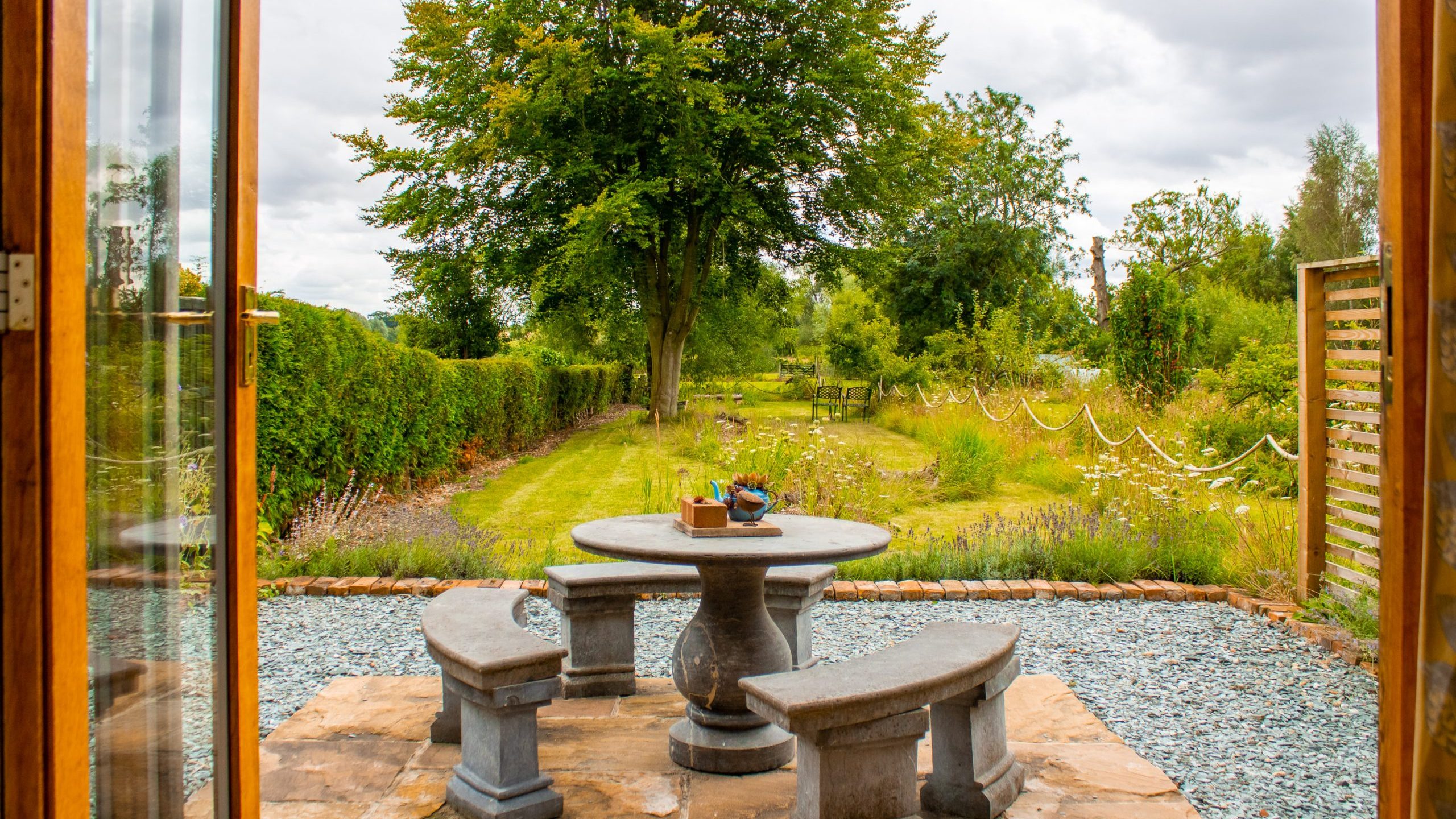 A serene garden view through open wooden doors, featuring a stone patio set with a round table and two curved benches. The garden is lush with greenery, trees, and a pathway bordered by flowers and shrubs, creating a tranquil outdoor space.
