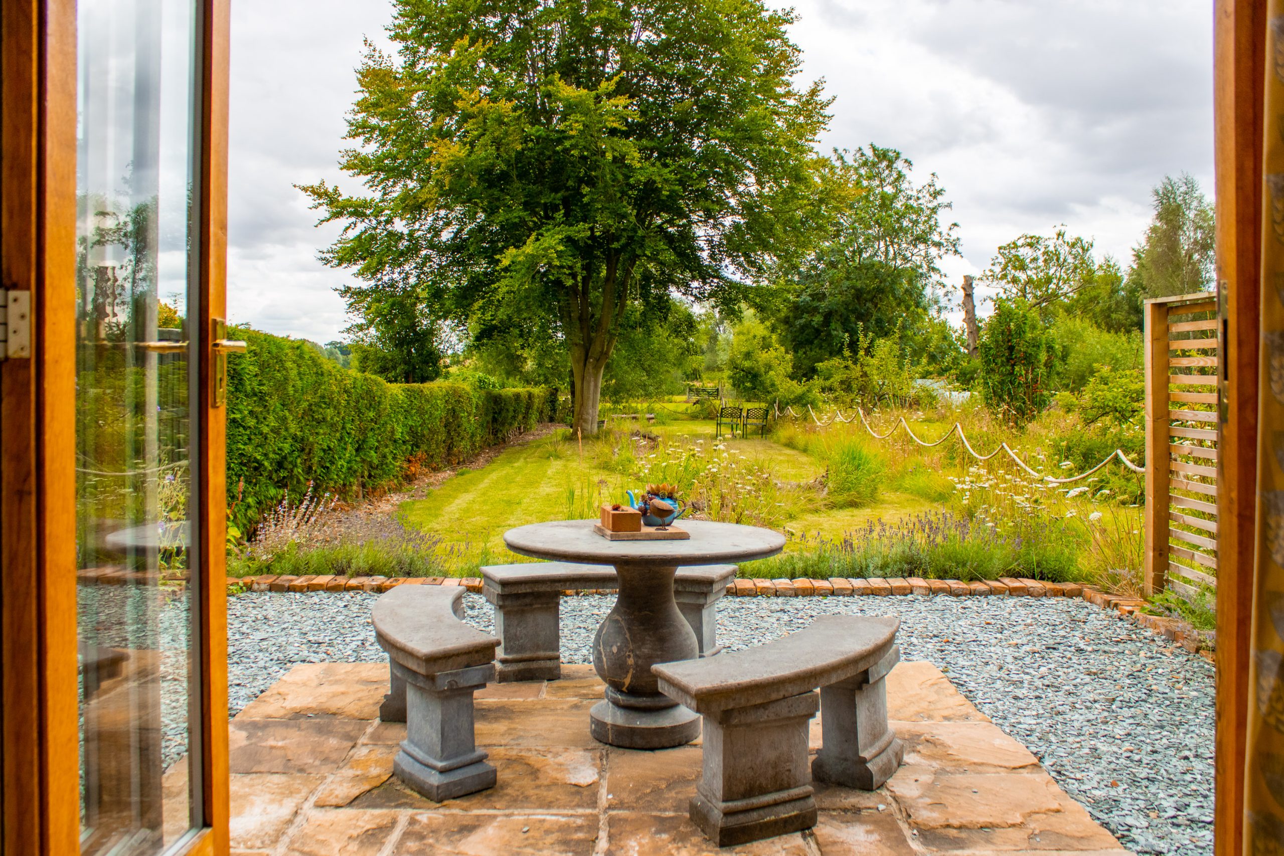 A serene garden view through open wooden doors, featuring a stone patio set with a round table and two curved benches. The garden is lush with greenery, trees, and a pathway bordered by flowers and shrubs, creating a tranquil outdoor space.