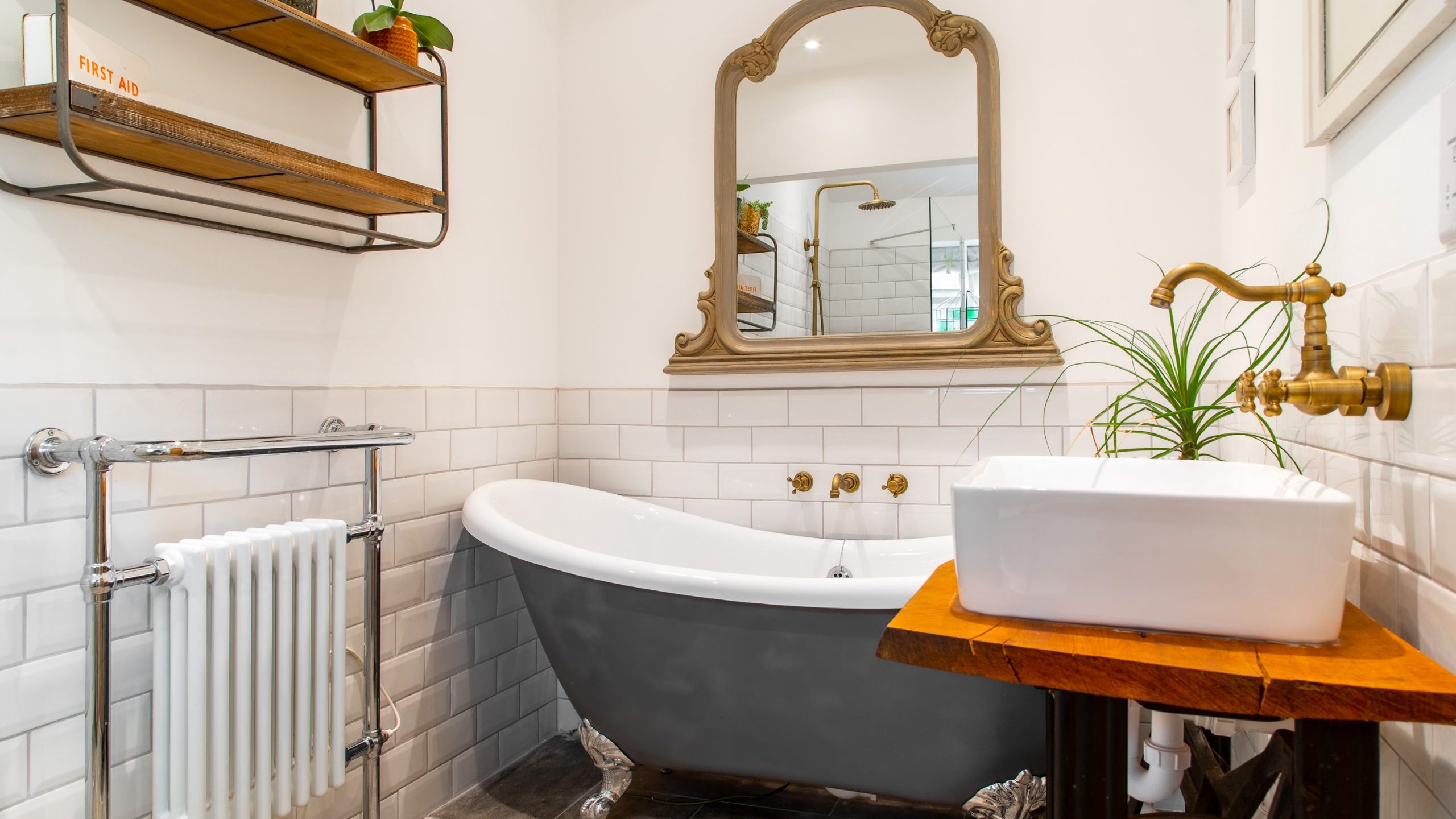 A cozy bathroom featuring a vintage clawfoot bathtub with silver feet and a freestanding faucet. A rectangular sink sits on a wooden vanity beside a potted plant. Above, a large mirror with a wooden frame is mounted on the wall. Shelves hold toiletries and decor.
