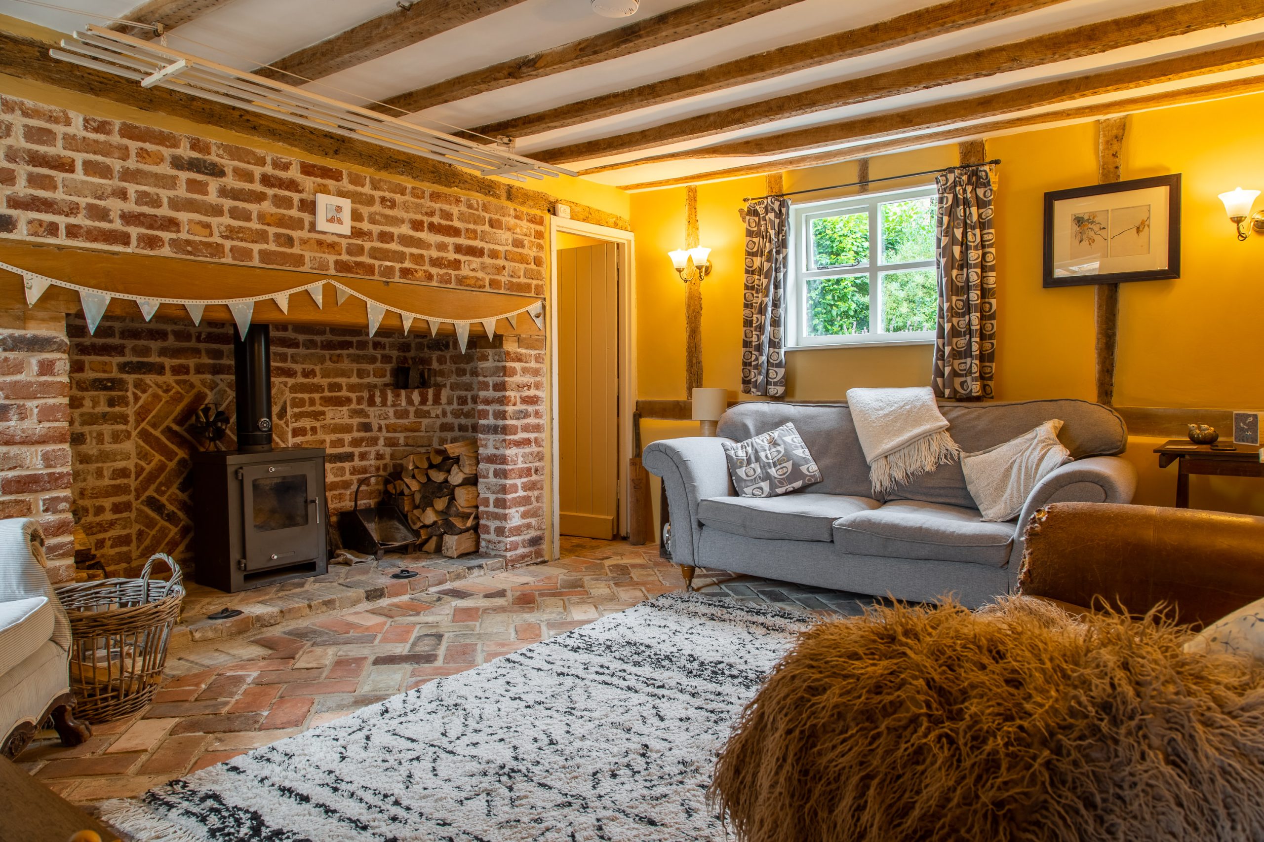A cozy living room with exposed wooden beams, a brick fireplace with a wood-burning stove, and a stack of firewood. The room features a gray sofa with cushions, a patterned rug, a window with curtains, a wall with warm yellow paint, and wall sconces for lighting.