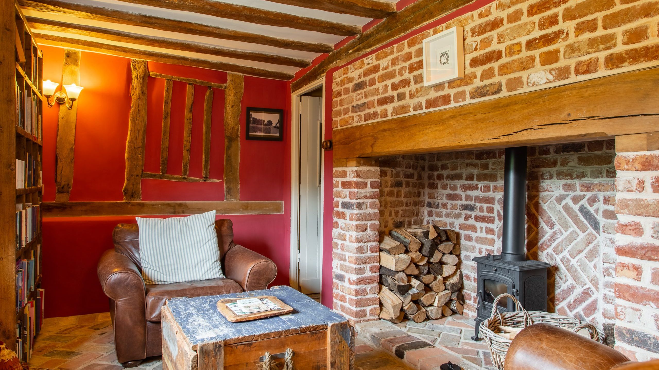 A cozy rustic room with exposed brick walls and wooden beams. Furnished with a leather armchair, a wooden chest used as a coffee table, and a log-burning stove with a stack of firewood. Walls are painted red, and a bookshelf is filled with books.