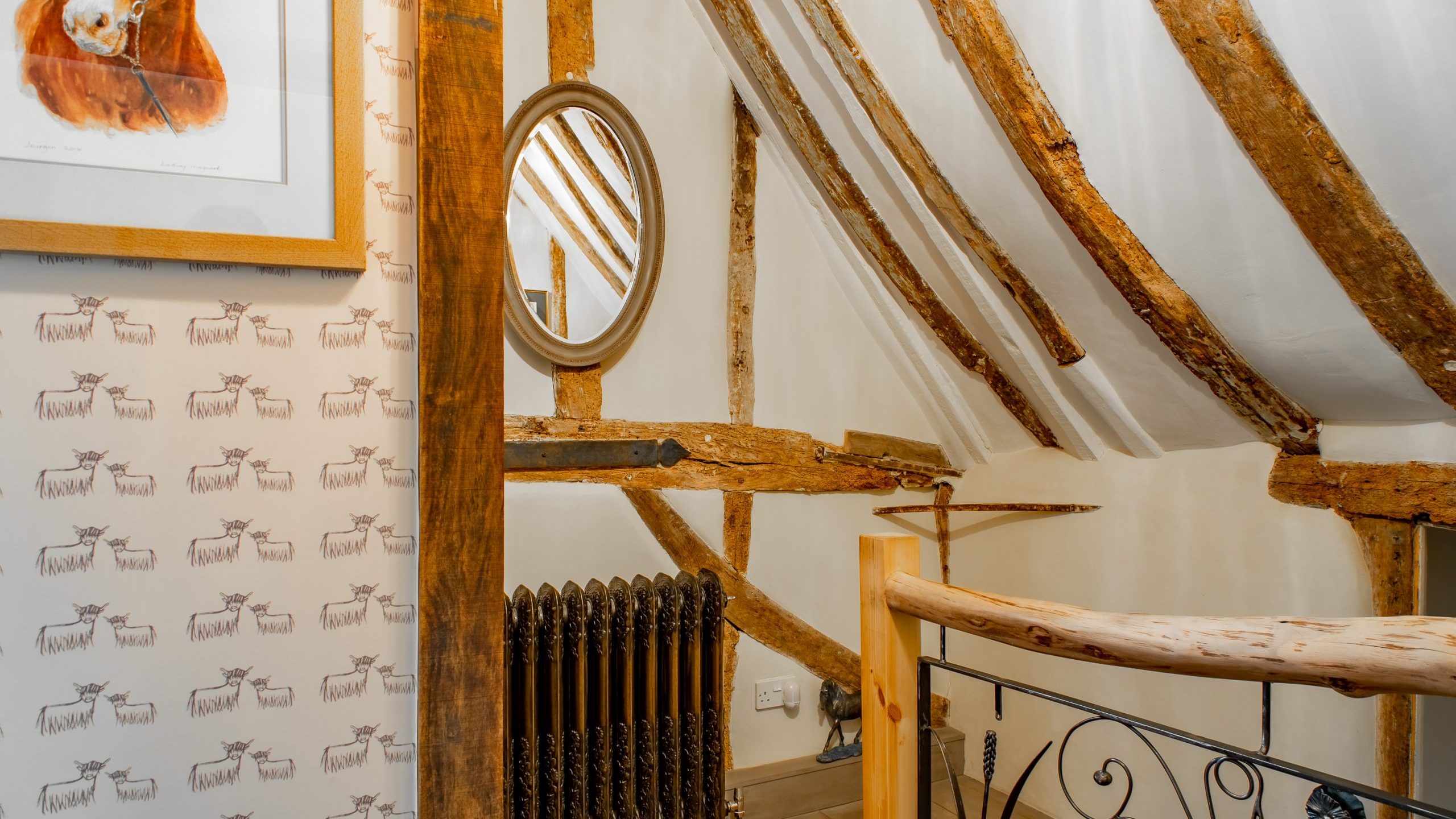A cozy room with exposed wooden beams on the ceiling and walls. A painting and a round mirror hang on one wall. The room features a decorative metal radiator cover and a rustic wooden bannister. The wall is adorned with patterned wallpaper.