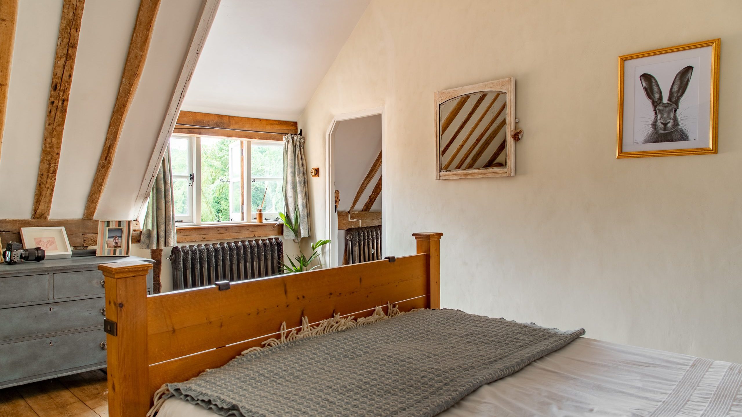 A cozy bedroom with a pitched ceiling featuring exposed wooden beams. The room includes a wooden bed with a gray blanket, a dresser with various items, a window with curtains, and framed artwork, including a rabbit picture, on the light-colored walls.