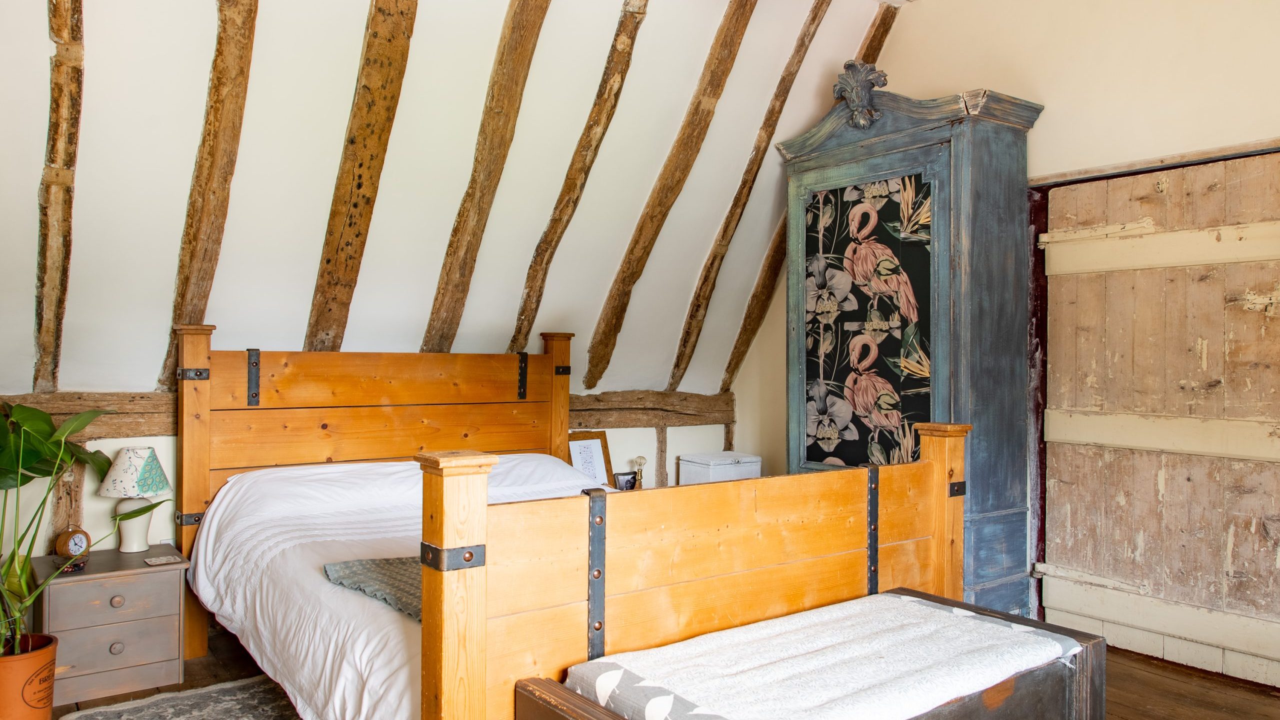 A rustic bedroom with exposed wooden beams on the slanted ceiling, a wooden bed frame, and white bedding. There is a large ornate blue wardrobe with floral design, a wooden chest at the bed's foot, nightstands, a potted plant, and a textured rug.