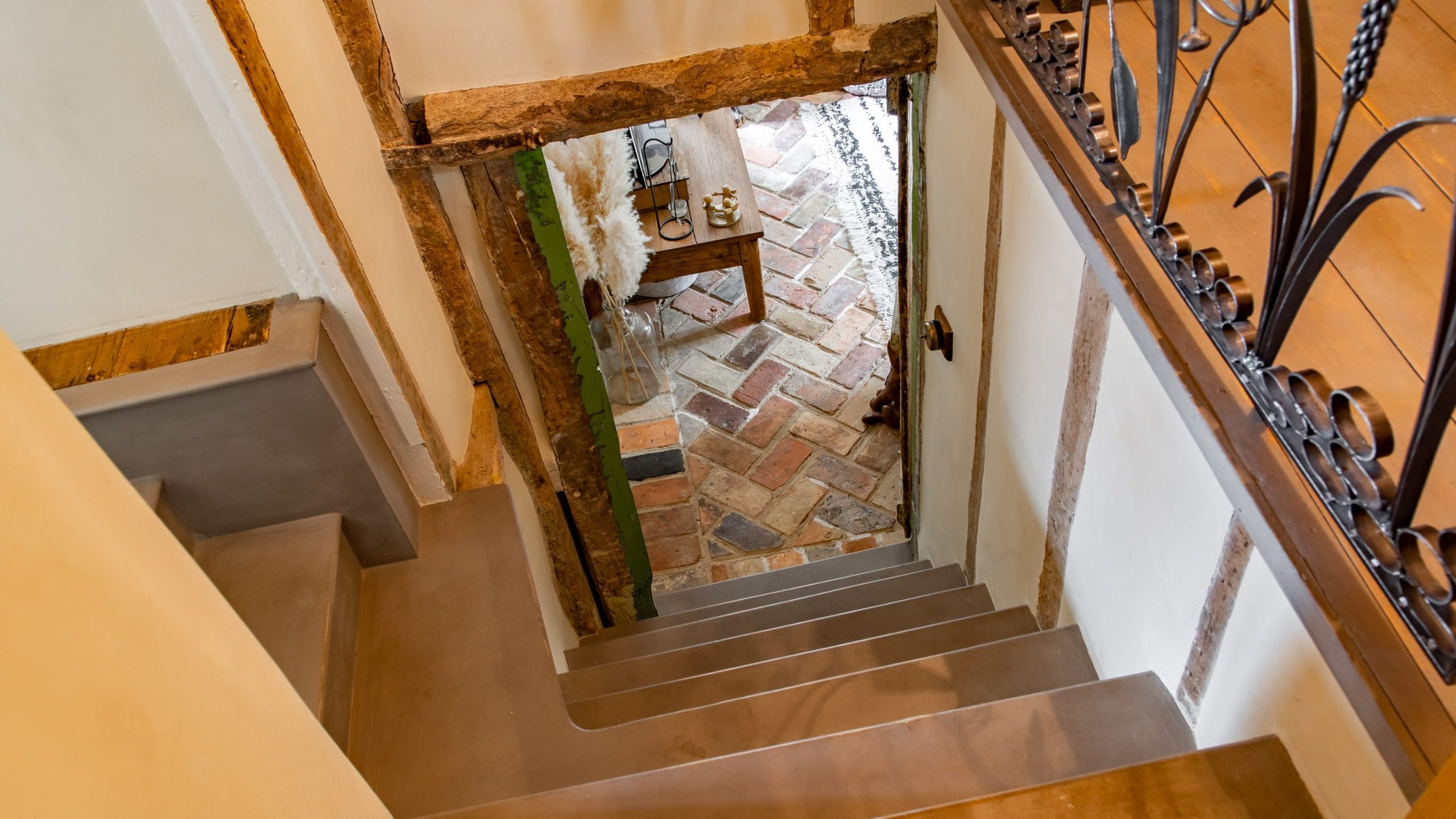 A view from the top of a narrow, steep staircase with stone steps leading down to a rustic hallway. The hallway features a patterned brick floor and decorative wrought iron railings. A small table with candles is visible through an open door at the bottom.