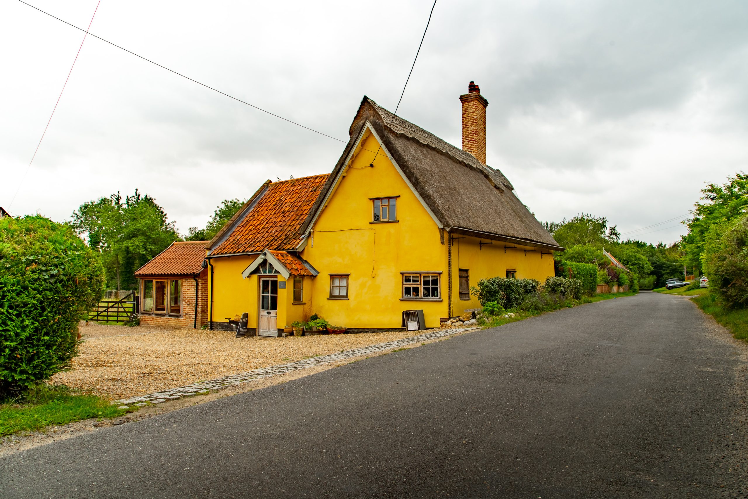 A charming yellow cottage with a thatched roof and a brick chimney stands beside a quiet country road. The cottage features white-framed windows and a small front porch. The area around the house is lush with greenery, and a gravel driveway is visible.
