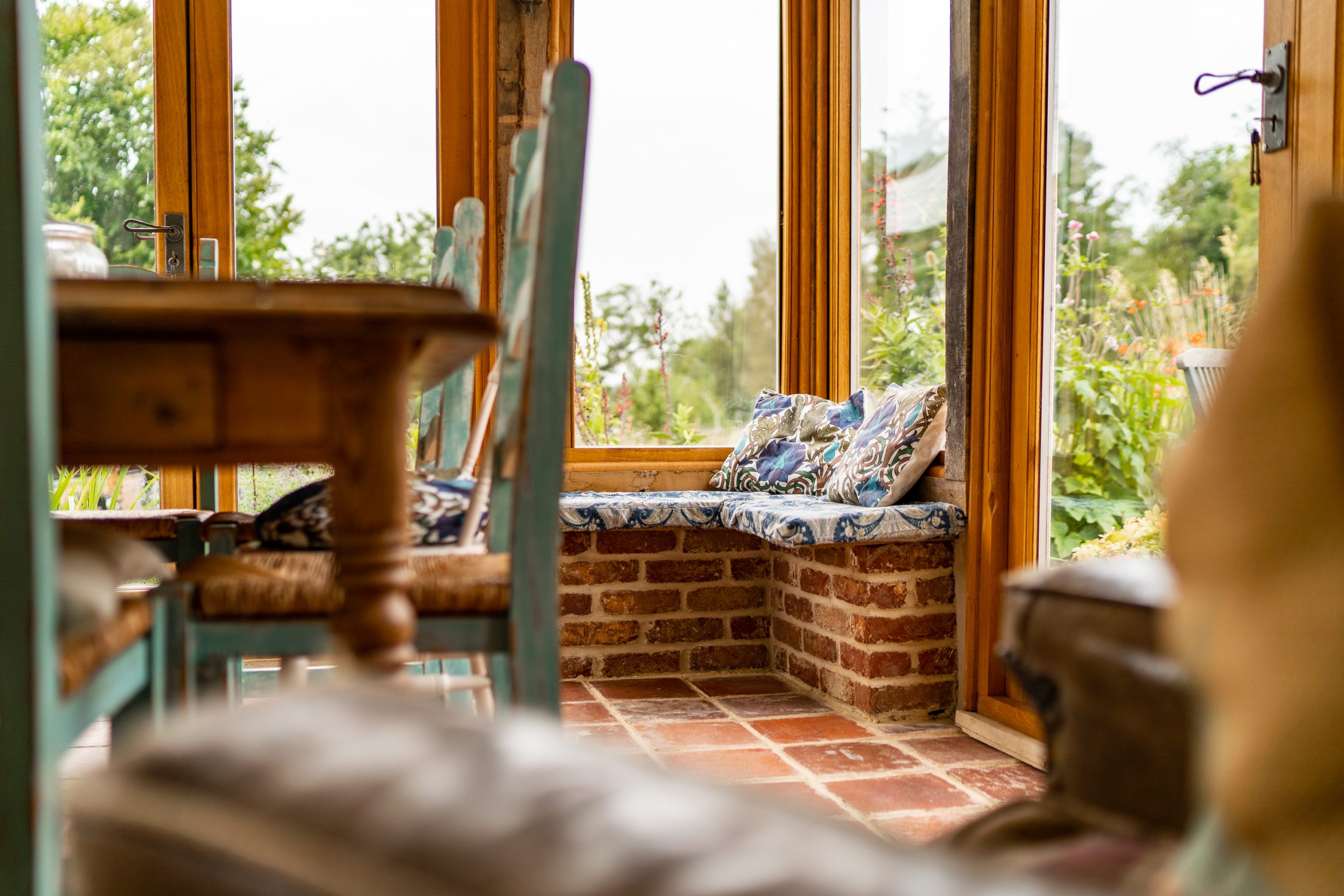 A cozy sunroom with large windows, brick accents, and wooden furniture. Cushioned benches are built into the brick wall, providing a comfortable seating area to enjoy the garden view outside. The tile flooring complements the warm, rustic ambiance of the space.