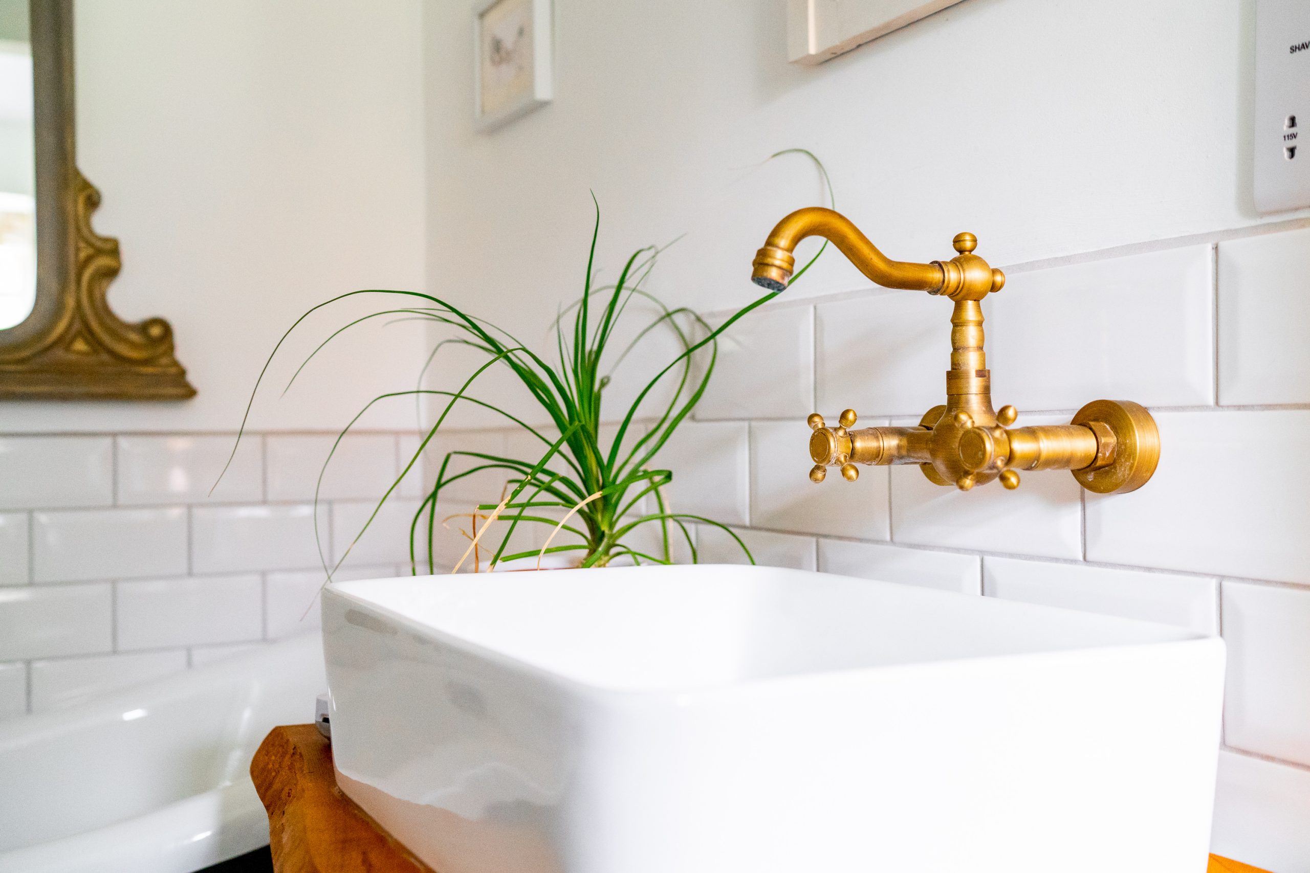 A modern bathroom features a white rectangular sink with a vintage-style brass faucet mounted on a white subway tile wall. A green potted plant adds a touch of nature. A partial view of a framed mirror with an ornate gold frame is visible on the left.