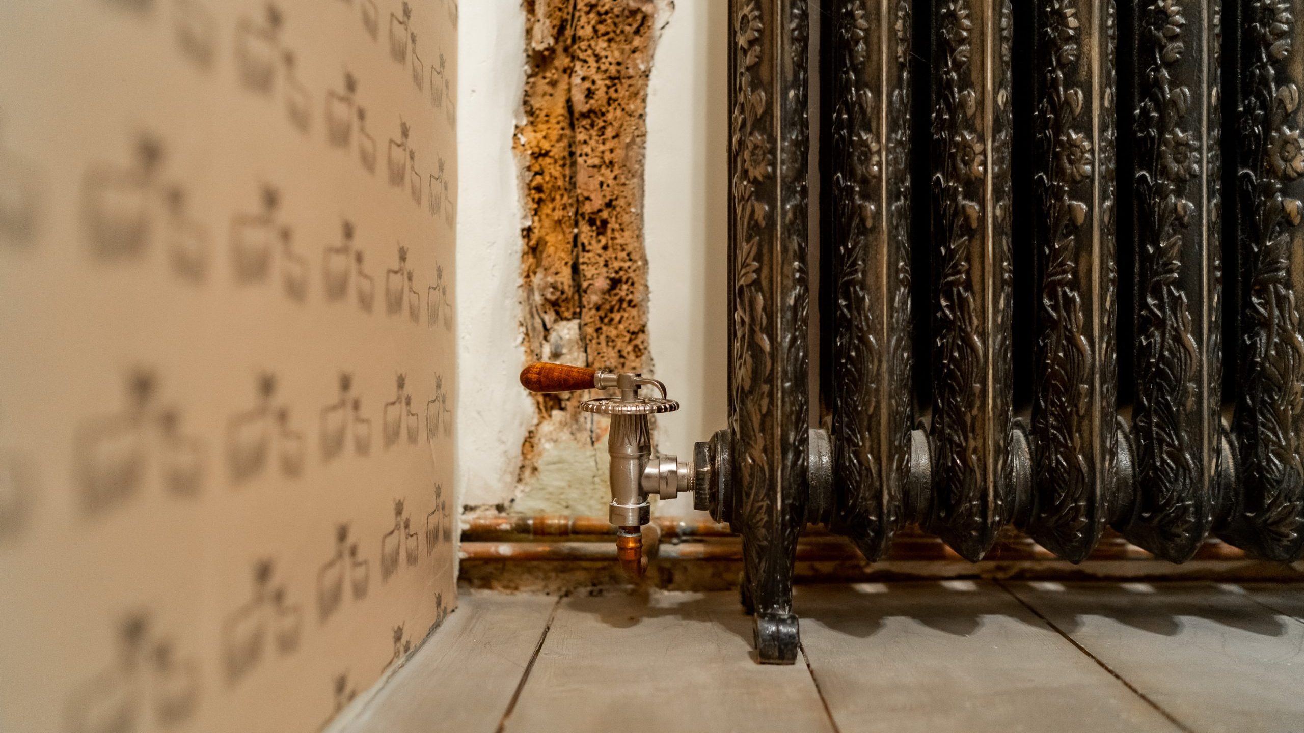 A close-up shot of an old, ornate cast iron radiator attached to a wall with peeling paint and exposed piping. The intricate design on the radiator stands out against the worn backdrop, while the wooden floor beneath adds to the rustic ambiance.
