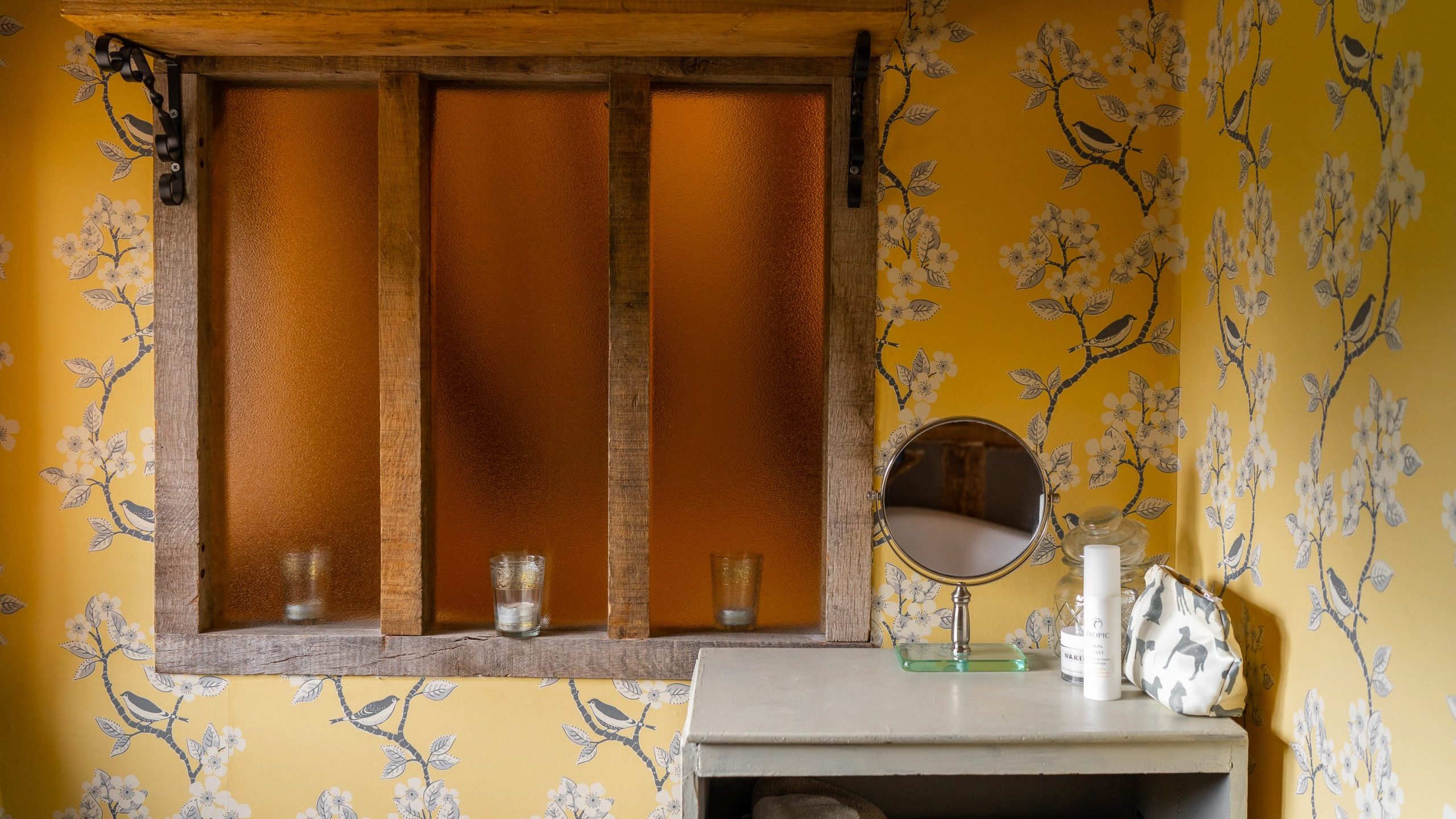 A cozy bathroom features yellow floral wallpaper, a wooden-framed translucent window, and a mirrored shelf. The shelf holds skincare products, a round vanity mirror, and neatly folded towels, creating a welcoming and organized space.
