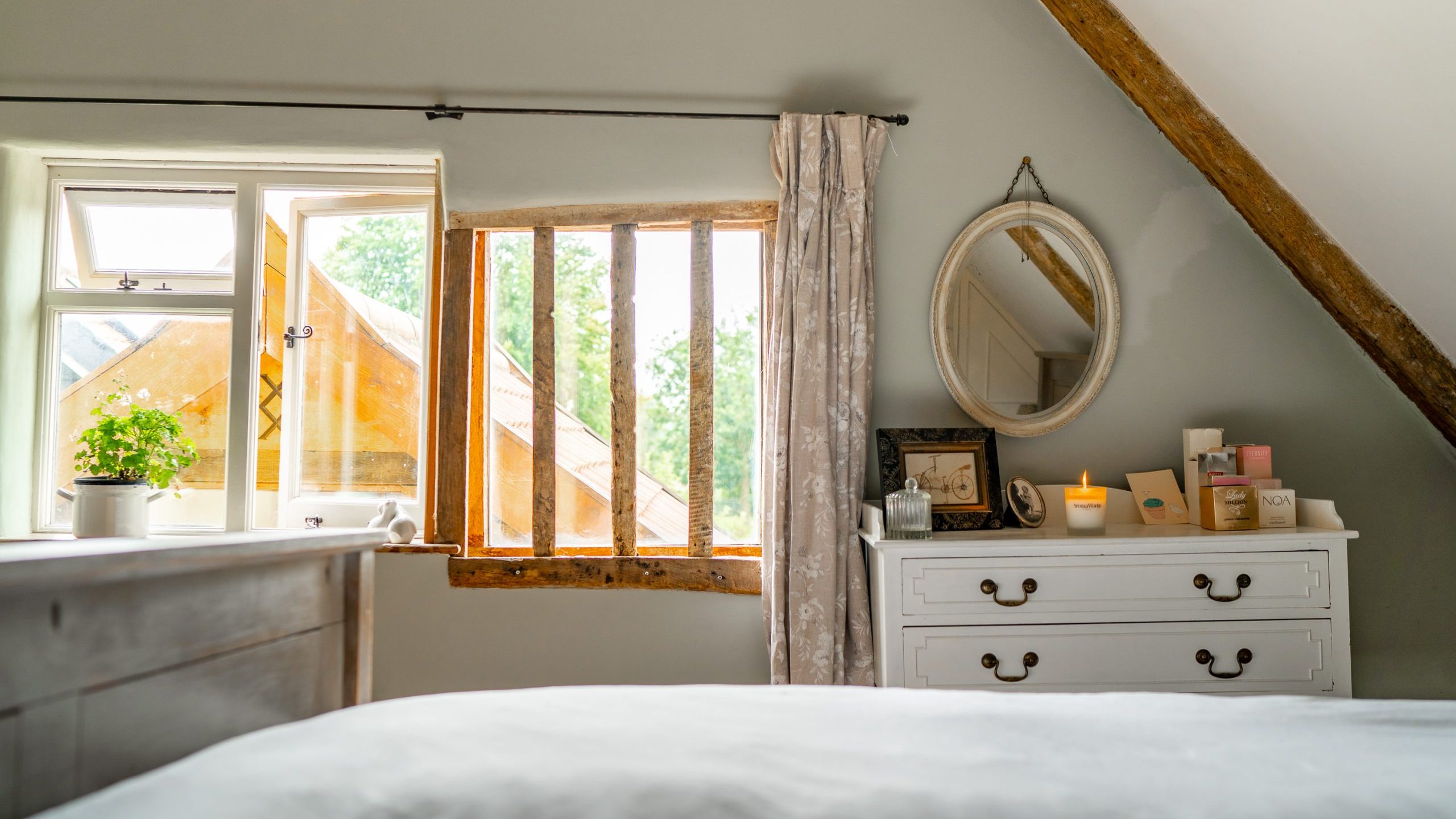 This cozy bedroom features a white dresser with candles, books, and a round mirror. A window with three panes reveals greenery outside. Another window is partially open, and a small potted plant sits on the sill. The room has sloped ceilings and wooden beams.