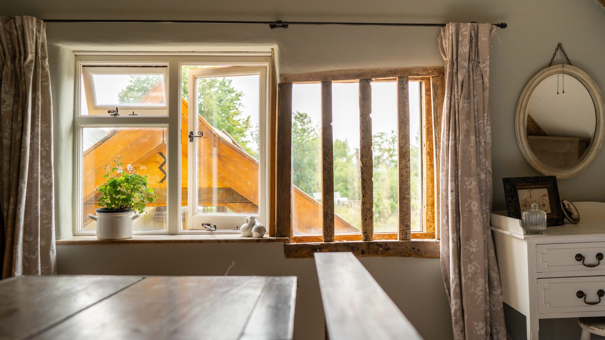 A cozy room with rustic decor features an unpainted wooden table in the foreground. Two windows with light curtains offer views of a green landscape and a wooden structure outside. A white dresser with framed photos and a round mirror adds to the room's charm.