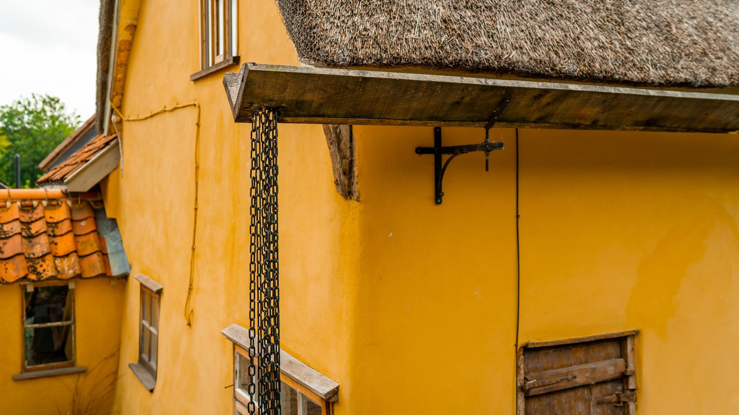 A close-up of a rustic yellow building with a thatched roof. The image shows a metal chain drainpipe attached to the building's corner, and an old wooden door below. The roof has wooden beams supporting it. The background includes greenery.